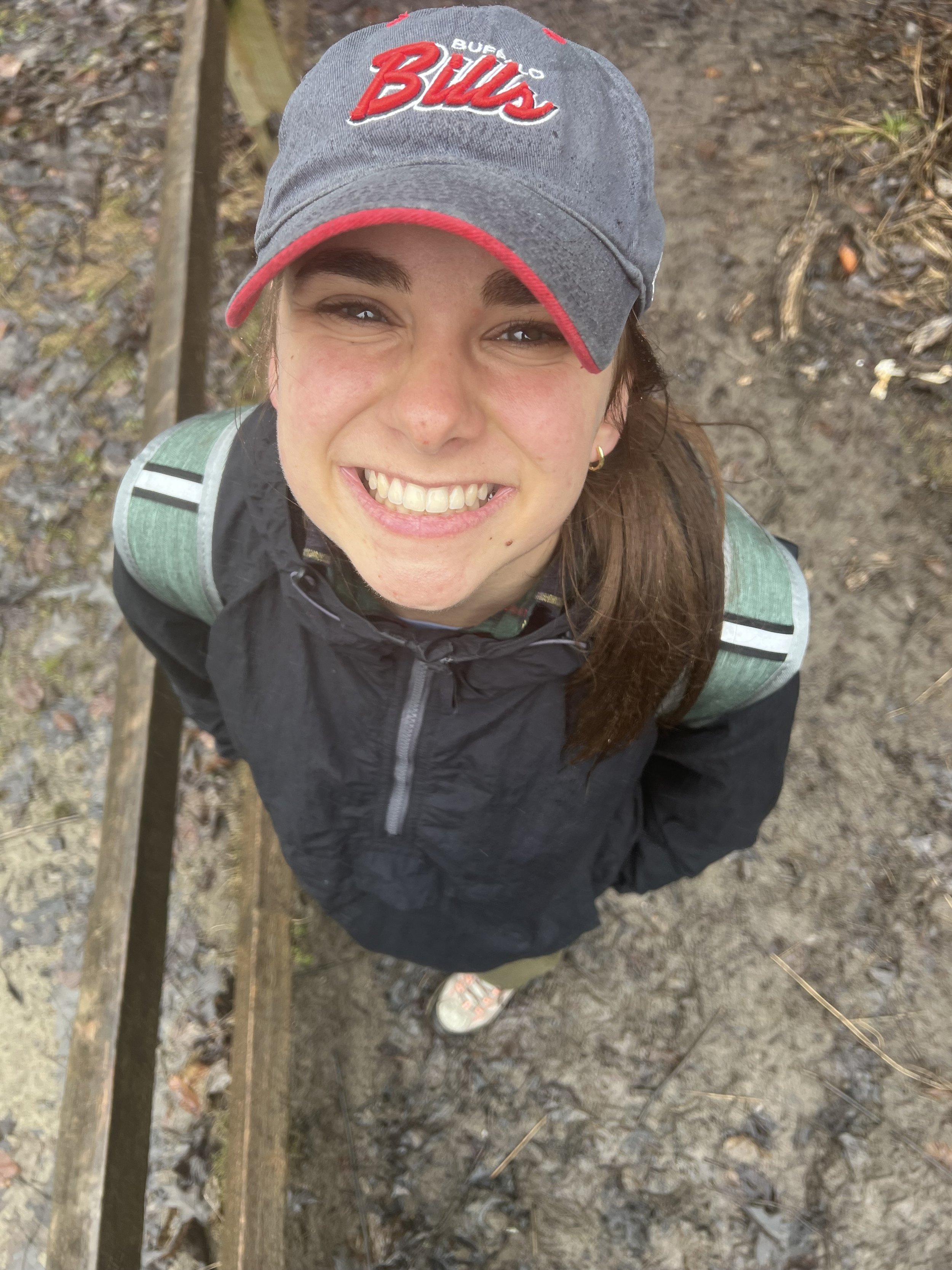 A young woman smiling and looking up at the camera while standing outdoors on a muddy trail. She is wearing a gray track cap with red accents and a black jacket, with a green backpack on her shoulders.