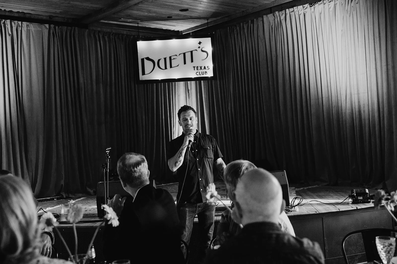 A speaker addressing dinner guests at Duetts, photographed in black and white.
