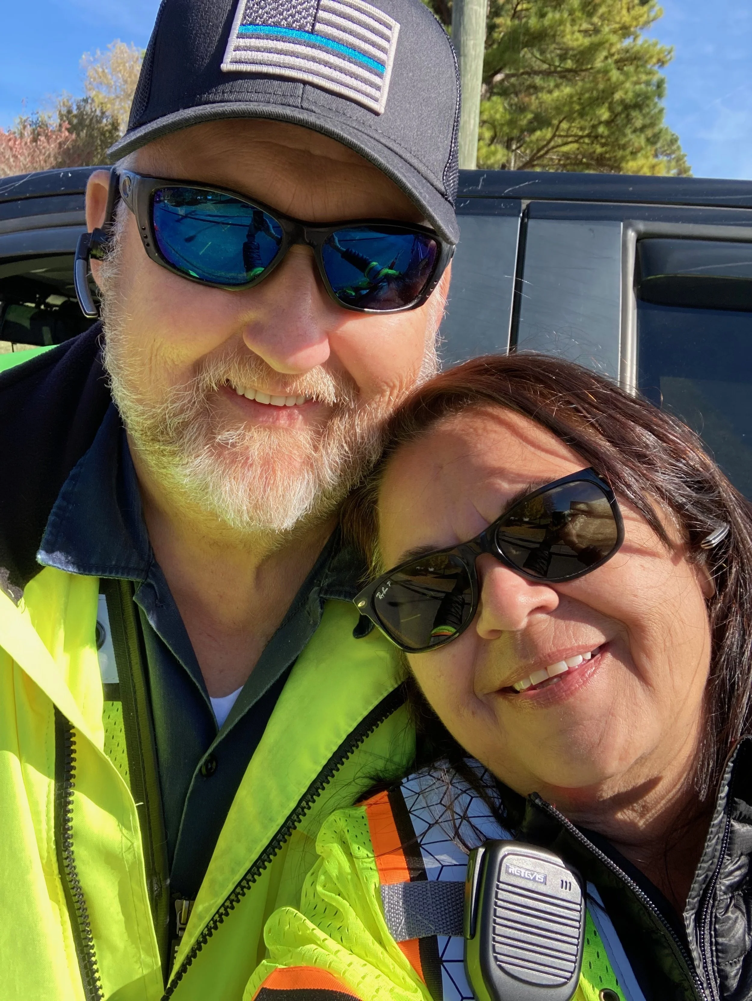 A smiling man and woman taking a selfie outdoors, both wearing sunglasses and yellow safety vests, with a vehicle and trees in the background.