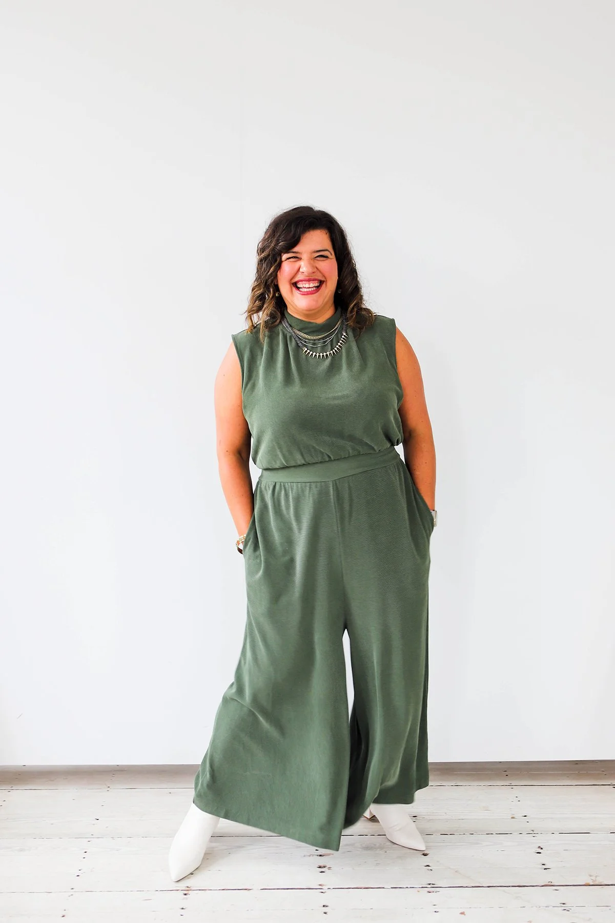 A woman with curly dark hair, wearing a green sleeveless top and matching wide-leg pants, stands smiling in front of a plain white wall.