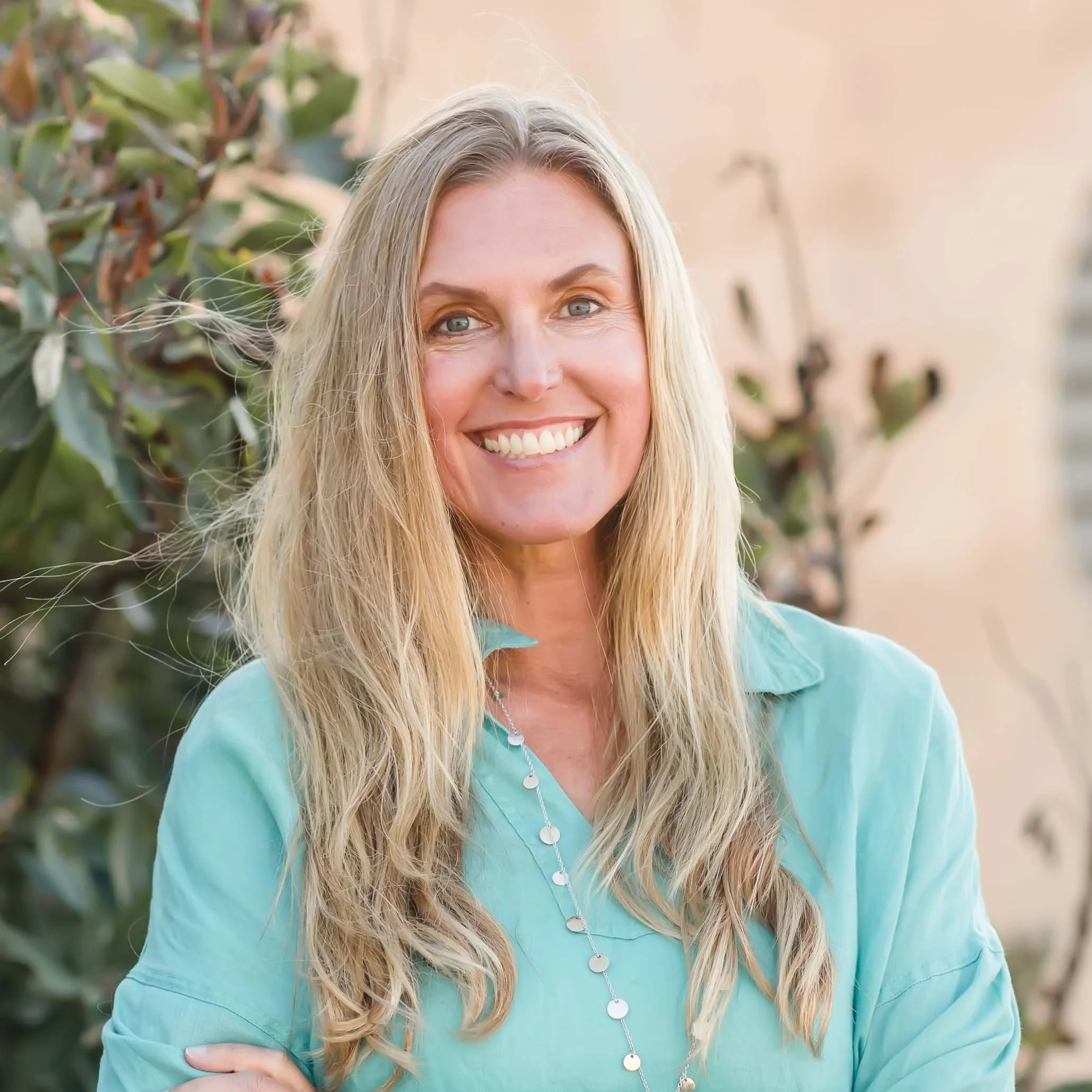 A woman with long blonde hair and a bright smile, wearing a light green shirt and standing outdoors with greenery in the background.
