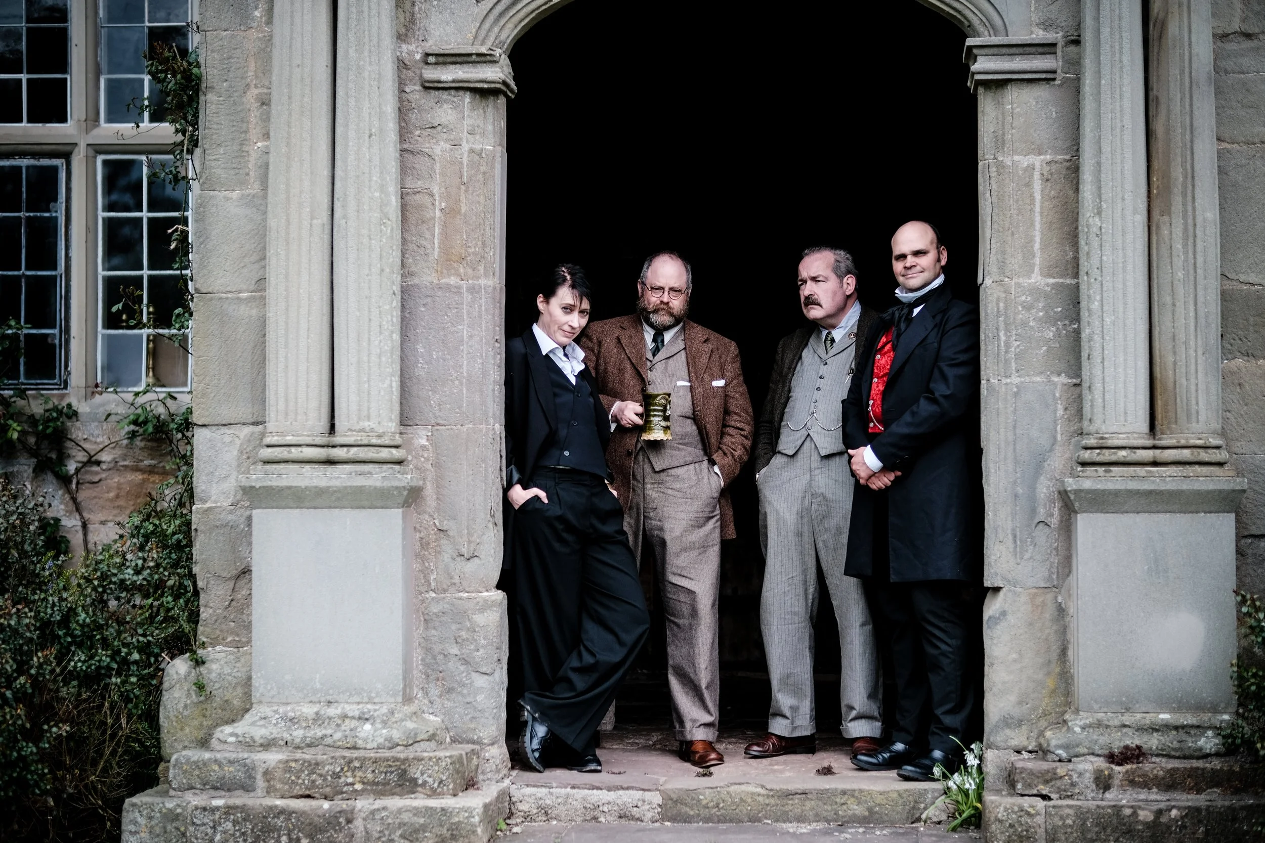 Four people dressed in vintage or formal attire stand in a stone archway of a historic building, with dark walls behind them. The group appears to be posing for a photo.