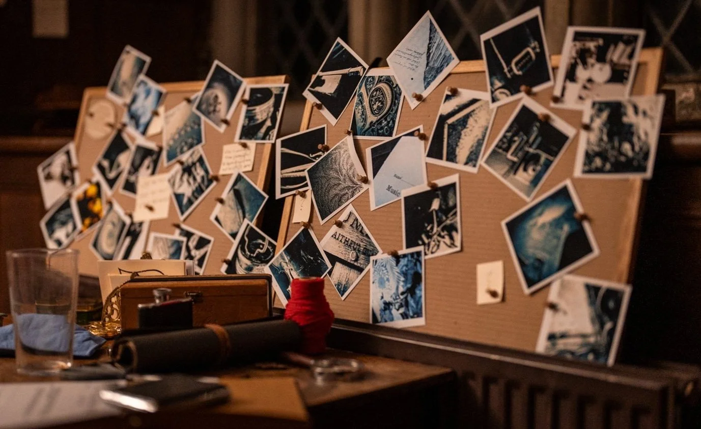 A bulletin board with numerous black and white photos and handwritten notes pinned to it, located on a dark wooden desk with various items including a glass of water, a rolled-up item, a red yarn spool, and some papers.