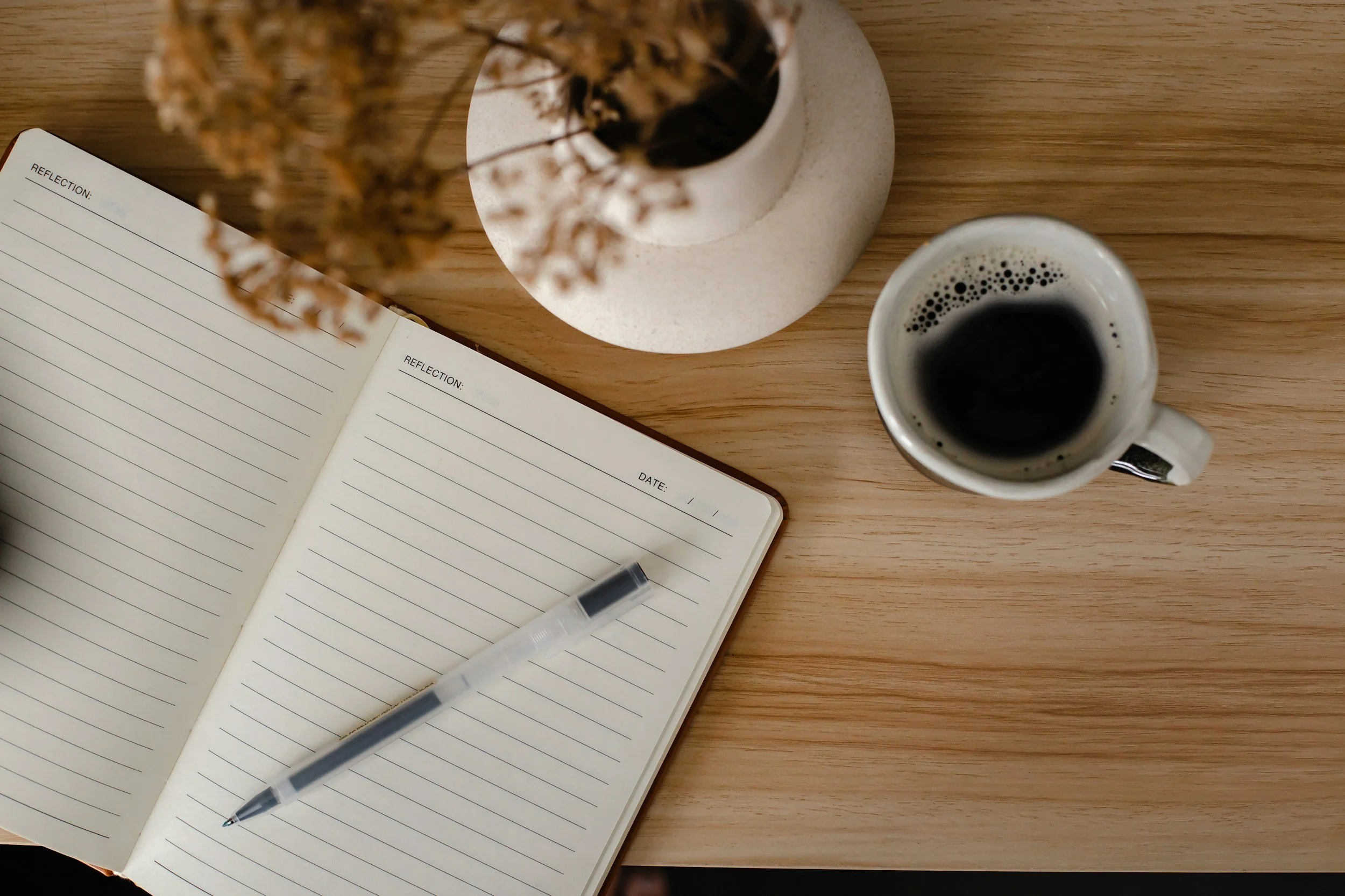 An open notebook with lined pages, a black pen on top, a white ceramic mug filled with black coffee, and a white vase with dried flowers on a wooden desk.