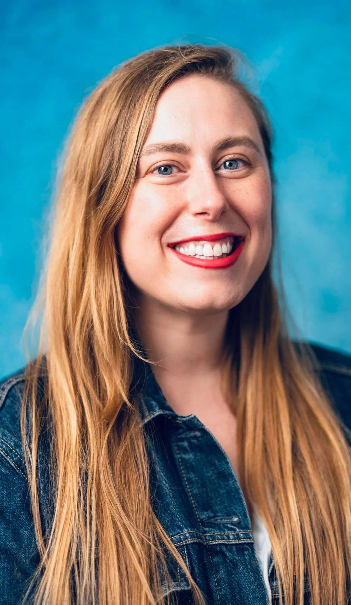 A smiling woman with long, red hair, blue eyes, red lipstick, and wearing a denim jacket against a blue background.