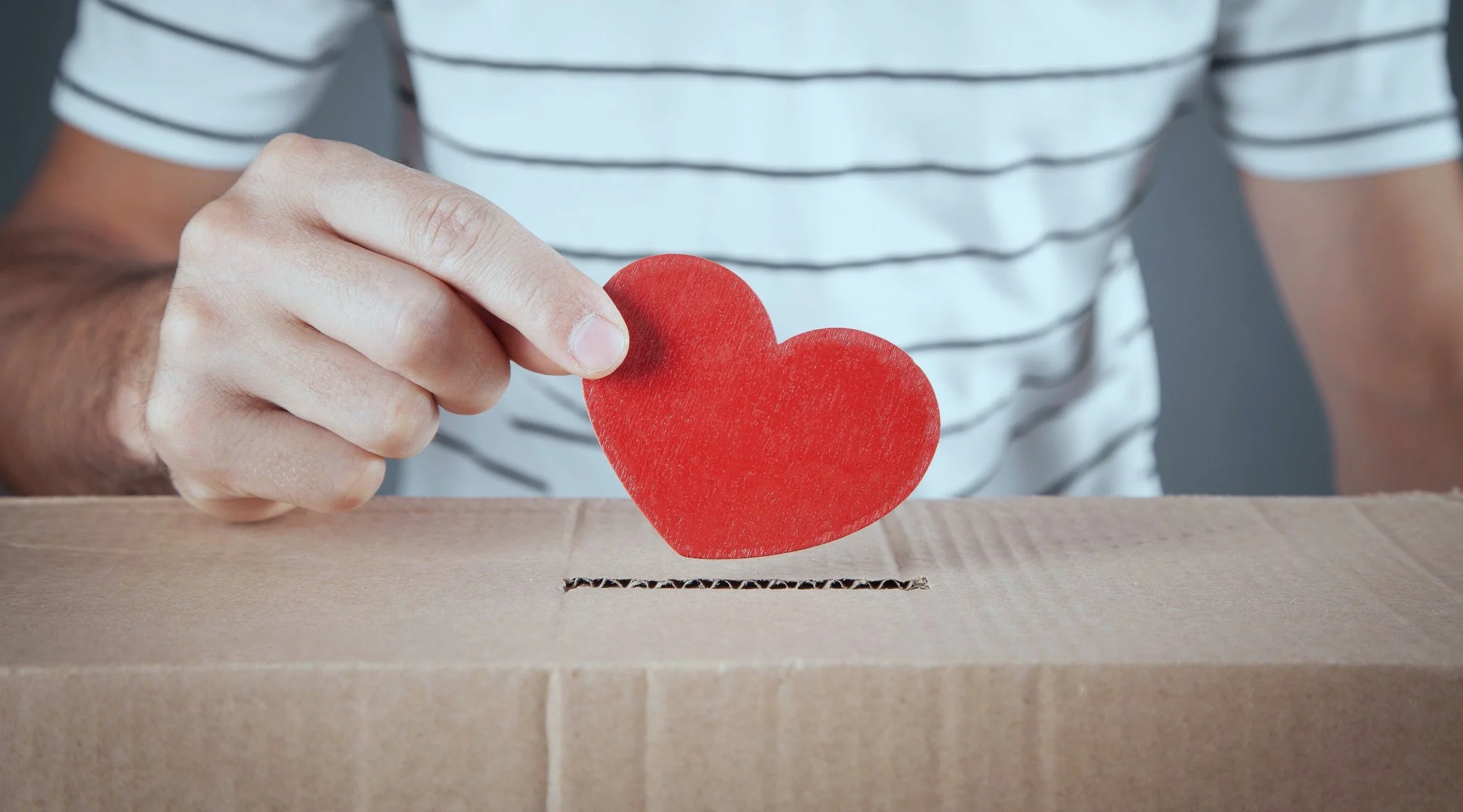 A person holding a red paper heart, preparing to put it into a cardboard box with a slit.