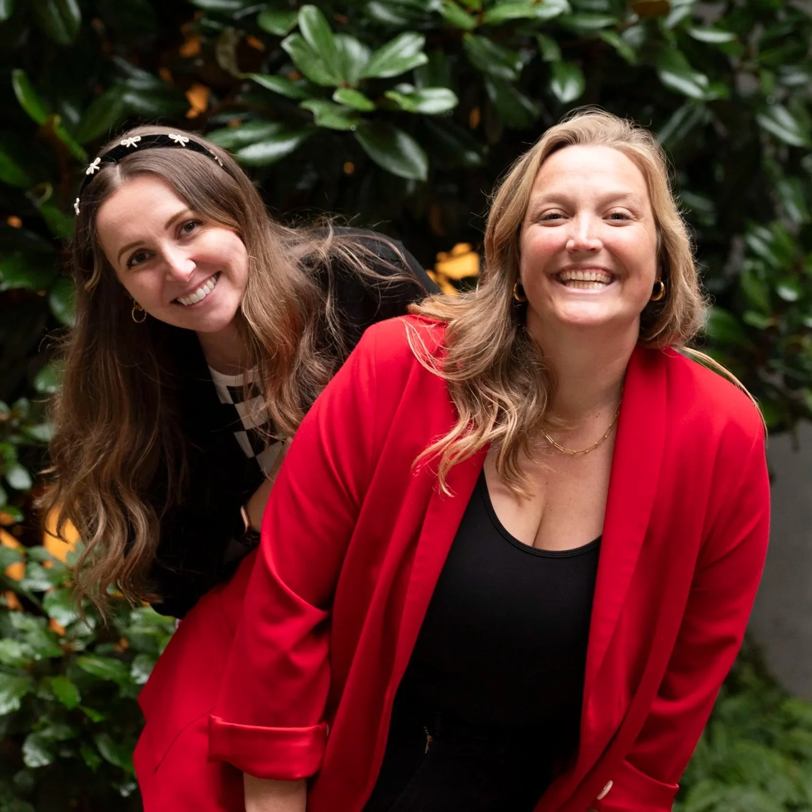 Mary with brown hair and black top standing behind Logan with blonde hair, red blazer and black top. Mary is leaning to the left and Logan is leaning to the right