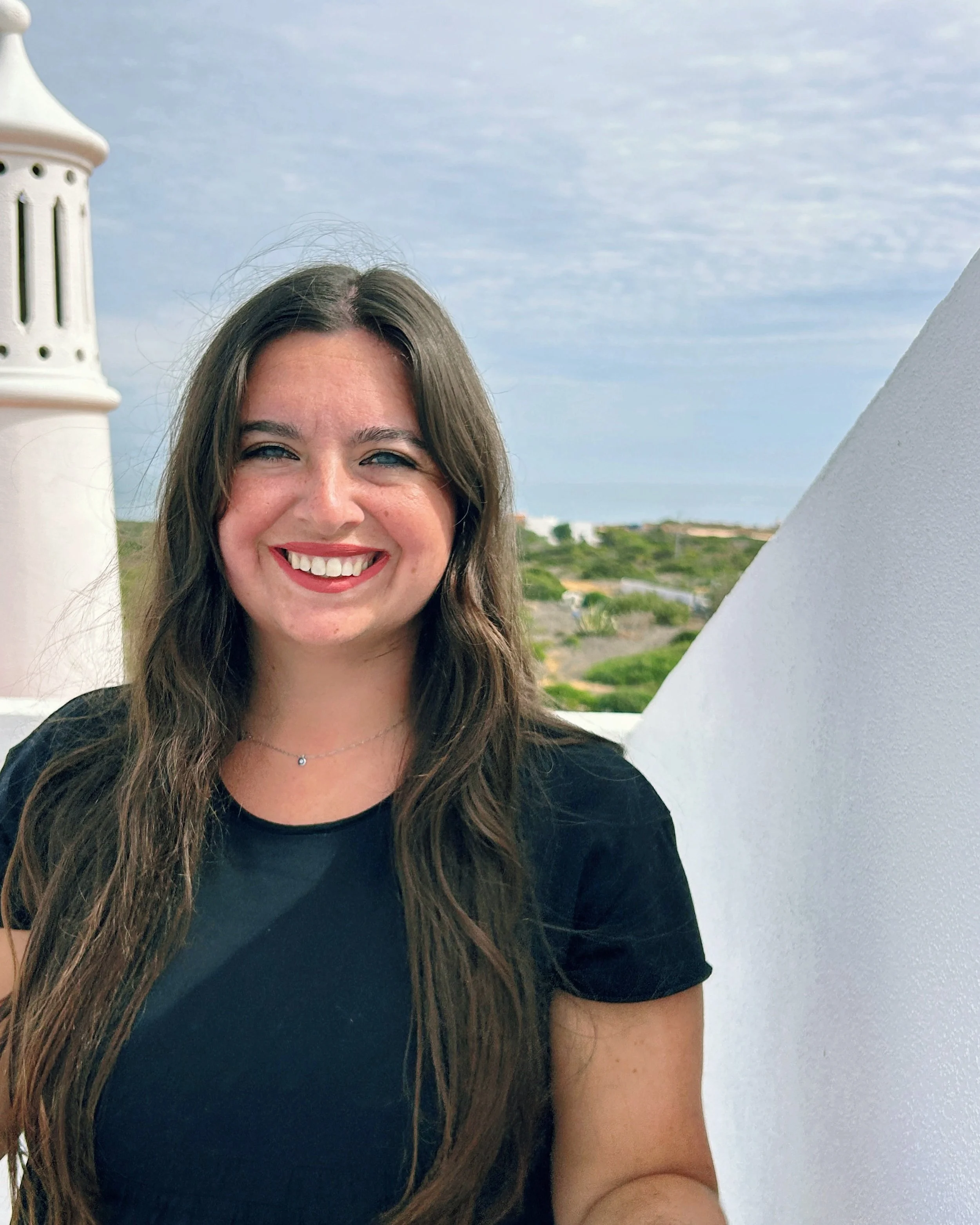 Dr. Sydney Conroy smiles to the camera with a sky and sea in the far background. She is on a rooftop with a smoke stack to her left and a slanted roof to the right. She is in a black dress with pink lipstick, her long brown hair down.