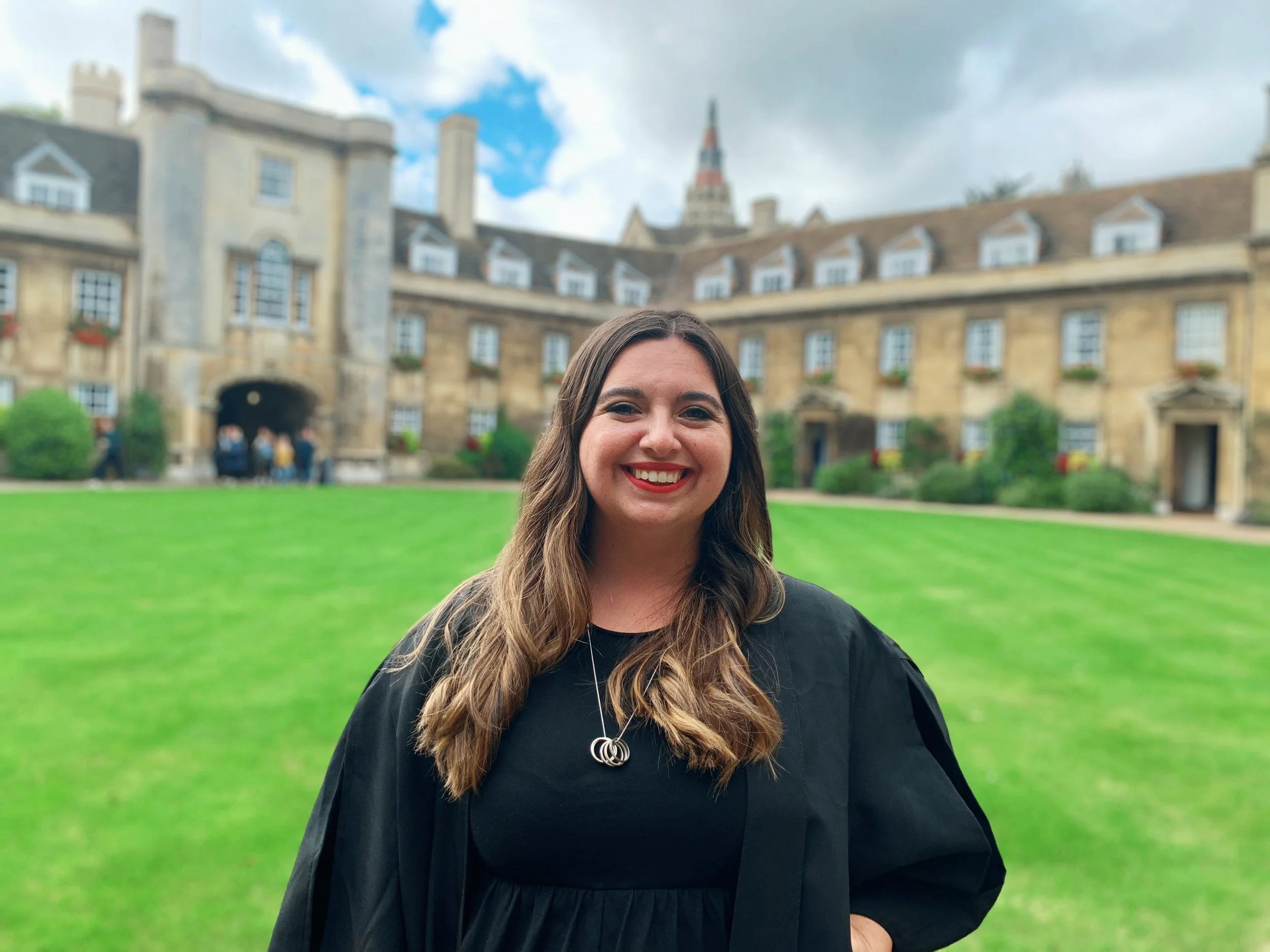 Sydney Conroy is standing up, smiling to camera with red lipstick, her dark brown hair is midlength and down. She is wearing a black dress with a silver necklace. She stands in the courtyard of Christs College at the University of Cambridge