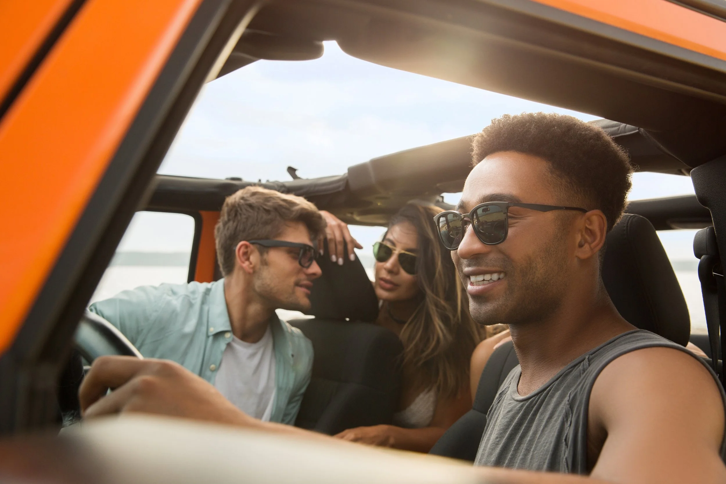 Four friends enjoying a drive in a convertible car on a sunny day, all wearing sunglasses and smiling.