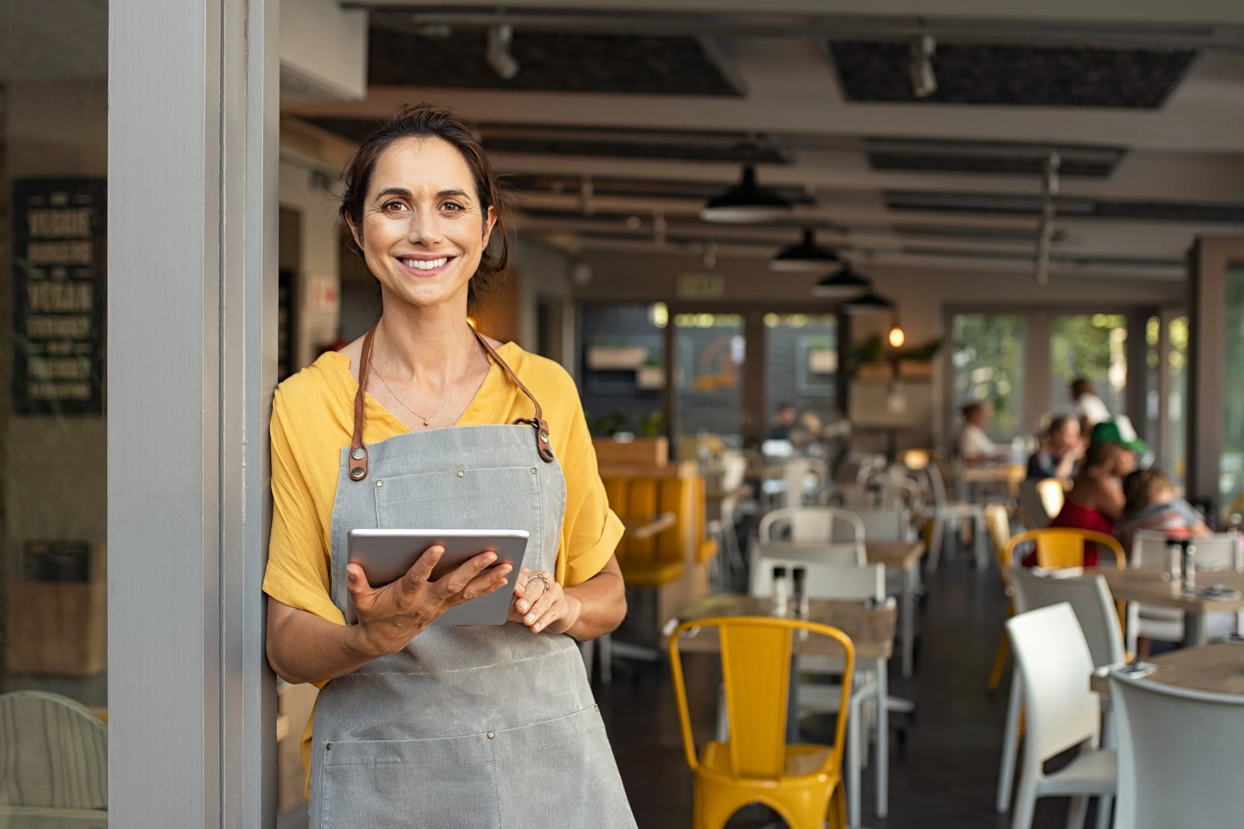 Smiling woman waitress in yellow shirt and gray apron standing at restaurant entrance, holding tablet.