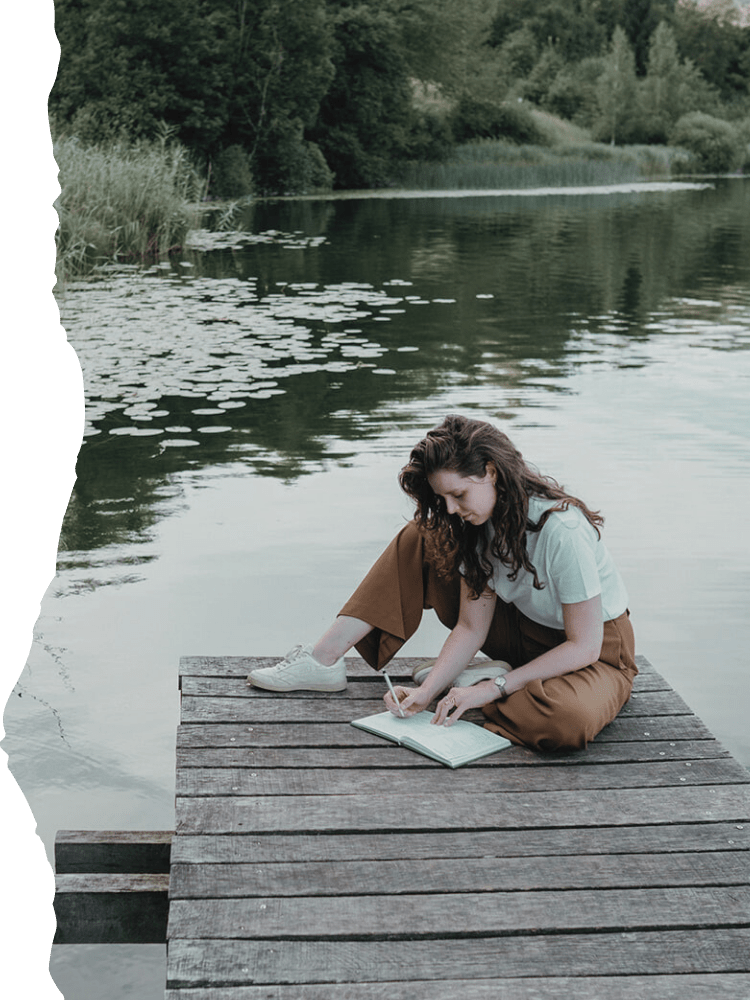 A young woman with long curly hair sitting cross-legged on a wooden dock by a lake, writing in a notebook with a pen, surrounded by a lush green landscape and water lilies.