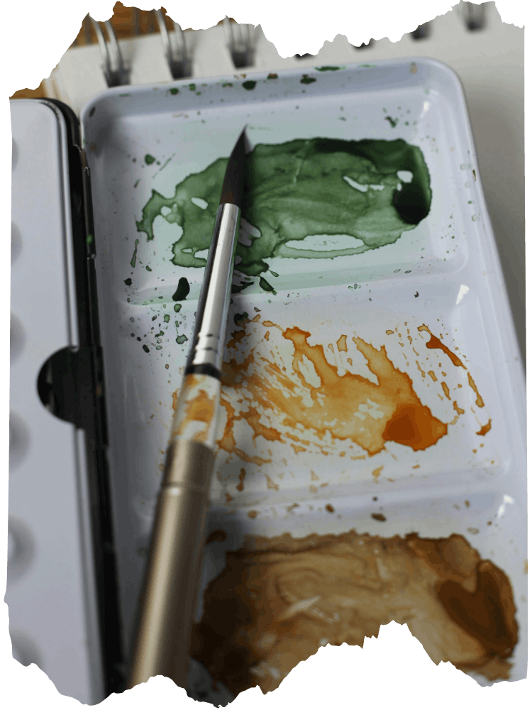 A paint palette with green, orange, and brown watercolor paint splotches, a silver paintbrush resting on it, and a blurred background.