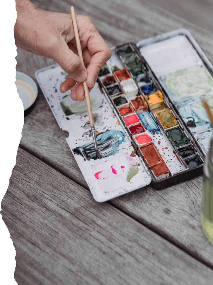 Close-up of an artist's hand holding a paintbrush, mixing blue watercolor paint on a white palette. The palette has various colors of watercolor paints arranged in compartments. The palette and paints are on a rustic wooden table.