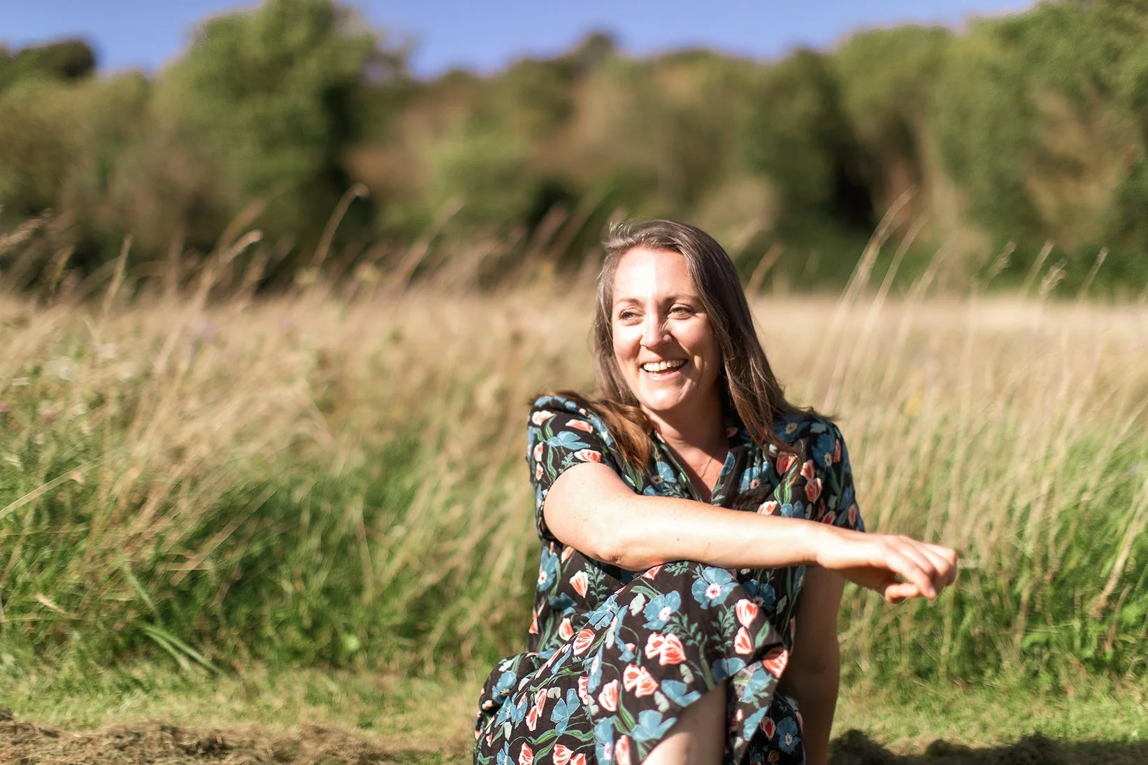 A woman sitting outdoors in a grassy field, smiling and wearing a floral dress.