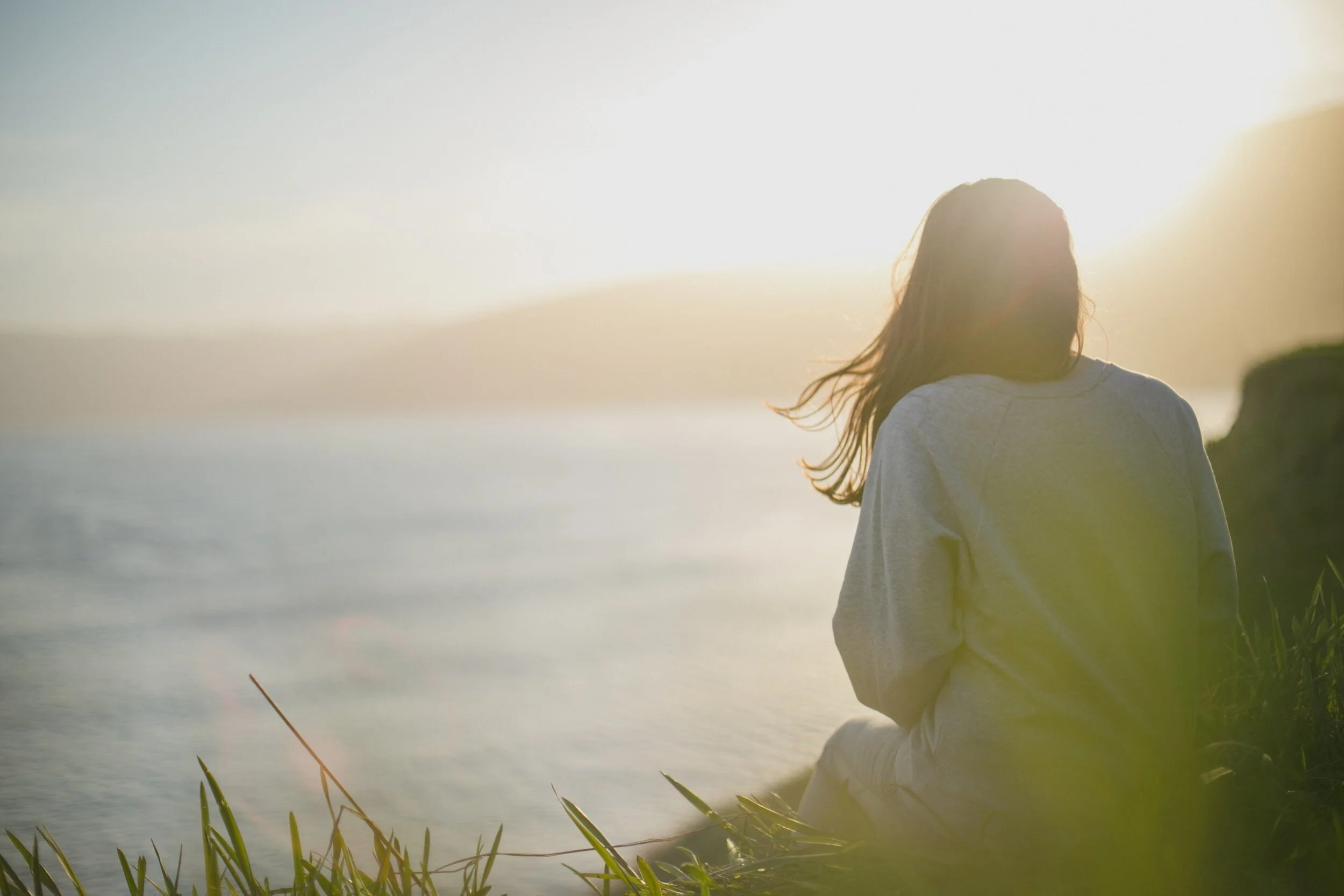 A person sitting on grass near water during sunset, facing away from camera.