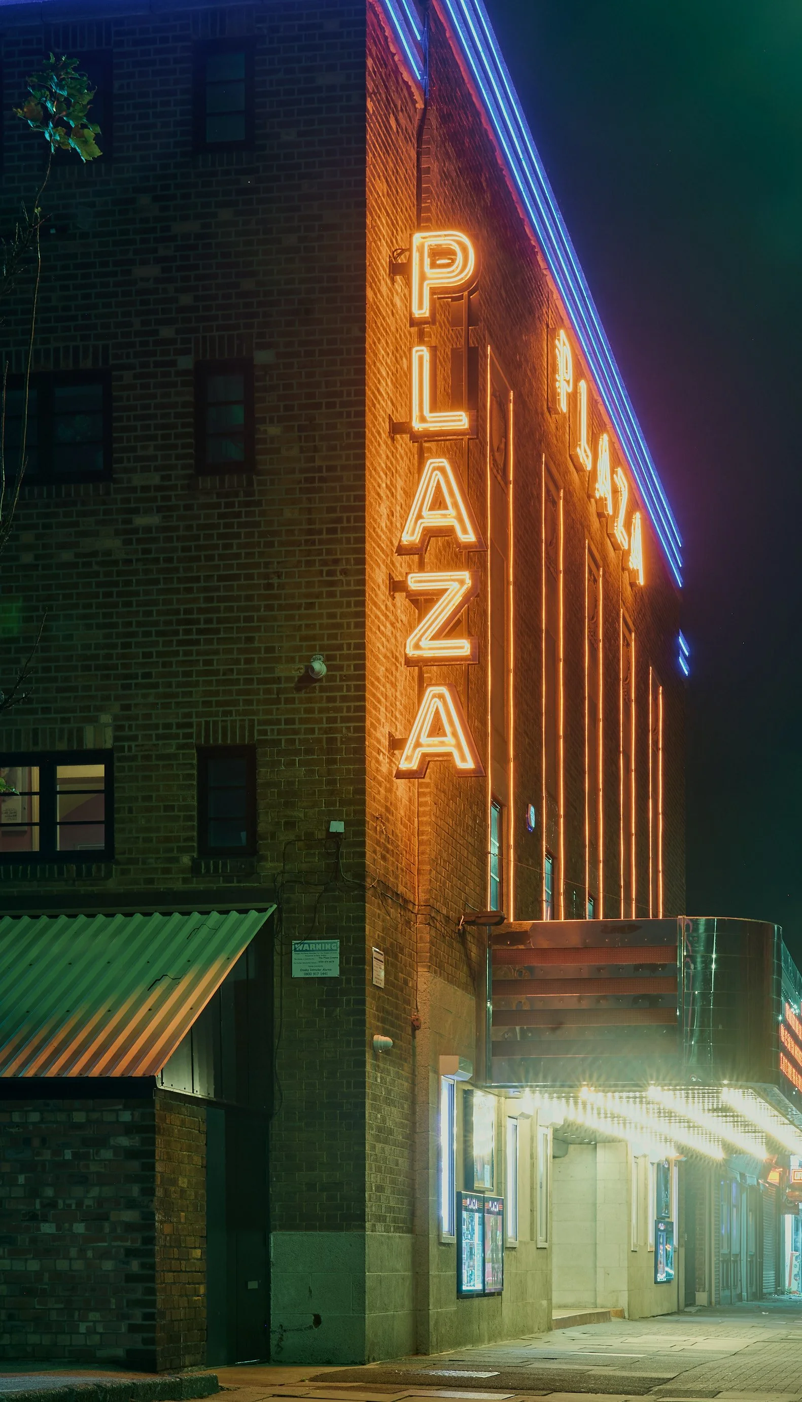 Neon sign reading 'PLAZA' on brick building at night with marquee and movie posters.