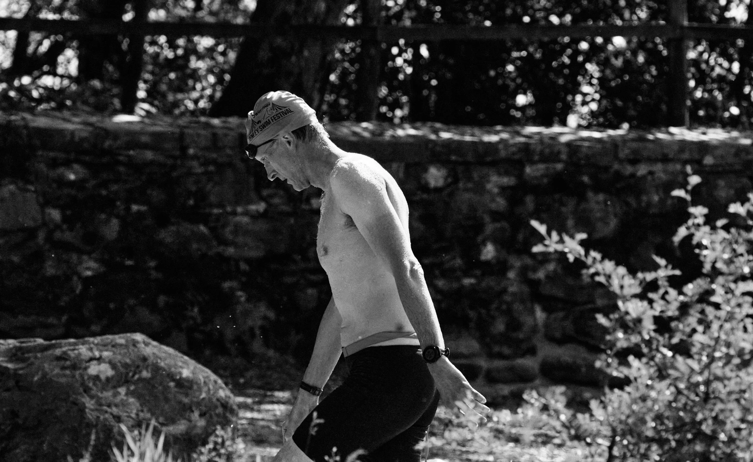 Black and white photo of a male swimmer in a cap and goggles walking on a rocky outdoor trail.