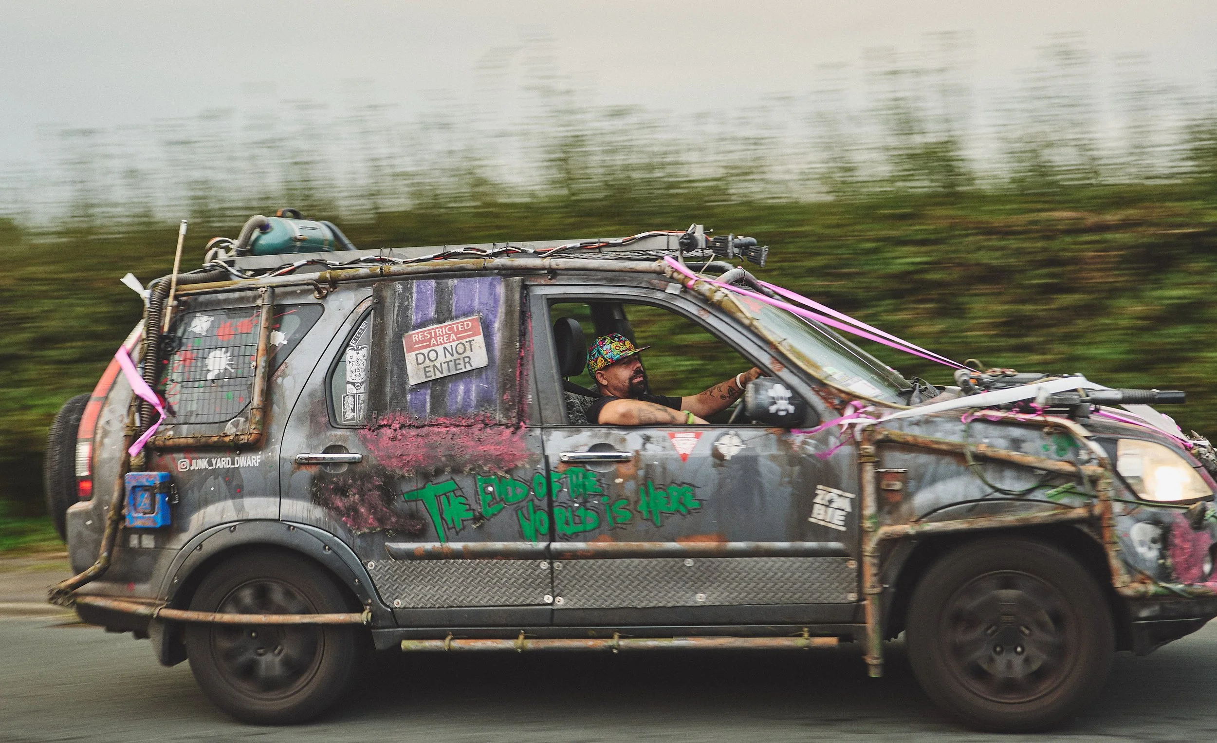 A heavily modified, graffiti-covered vehicle with a man driving, wearing a colorful hat. The vehicle has signs like 'RESTRICTED AREA DO NOT ENTER' and 'THE EDGE OF THE WORLD IS HERE,' with various decorations and attachments, in motion with a blurred