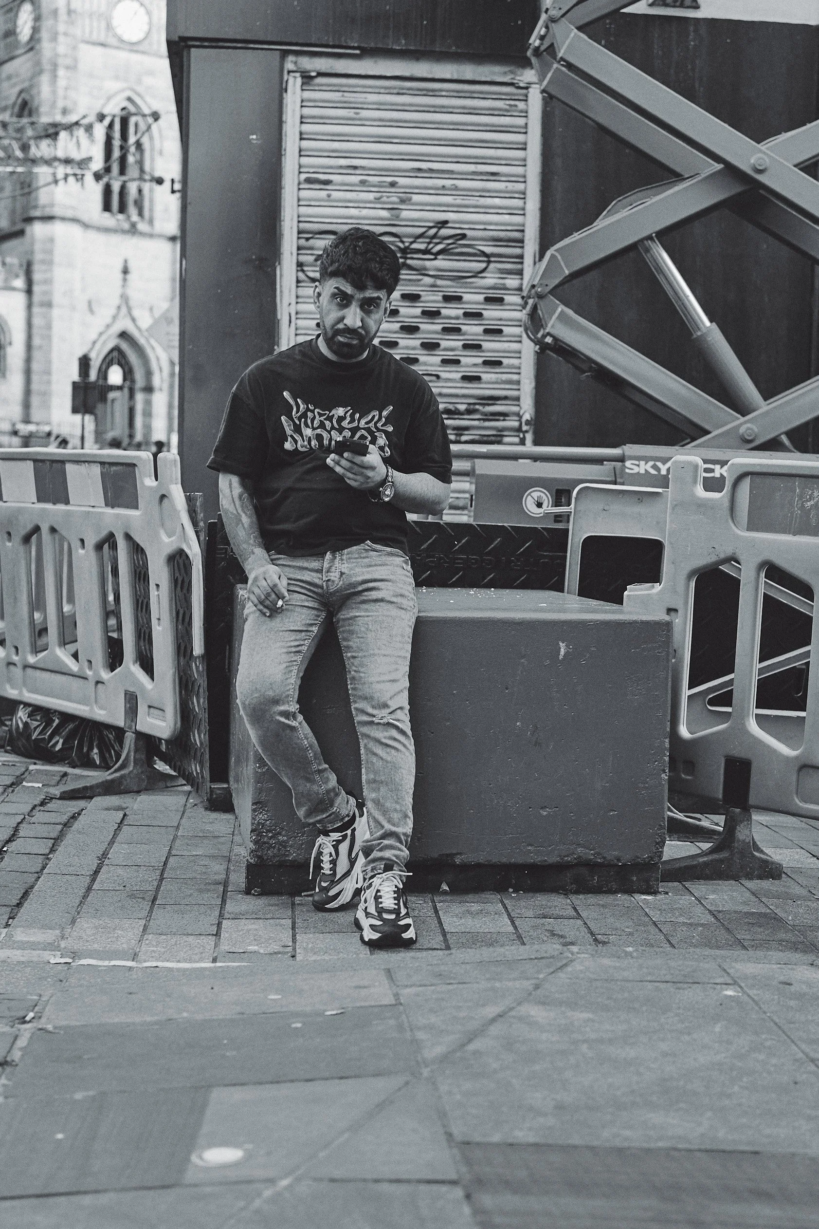 A man with a beard and tattooed arm in casual clothing leaning against a concrete barrier on a city street, looking at his phone, with scaffolding and building in the background.