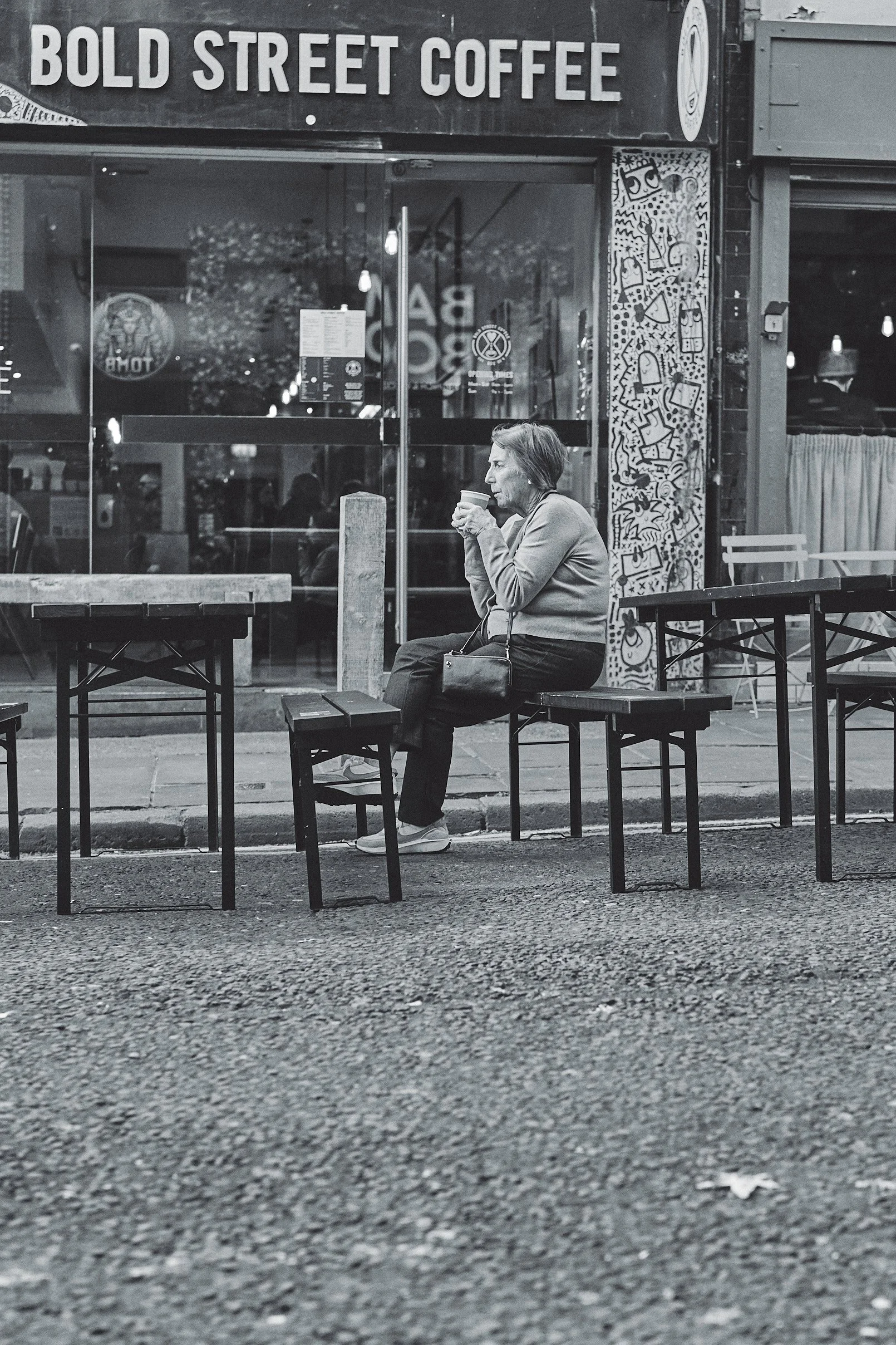 A woman sitting outside a coffee shop, drinking from a cup, with empty tables and chairs around her and the shop sign reading 'BOLD STREET COFFEE' in the background.