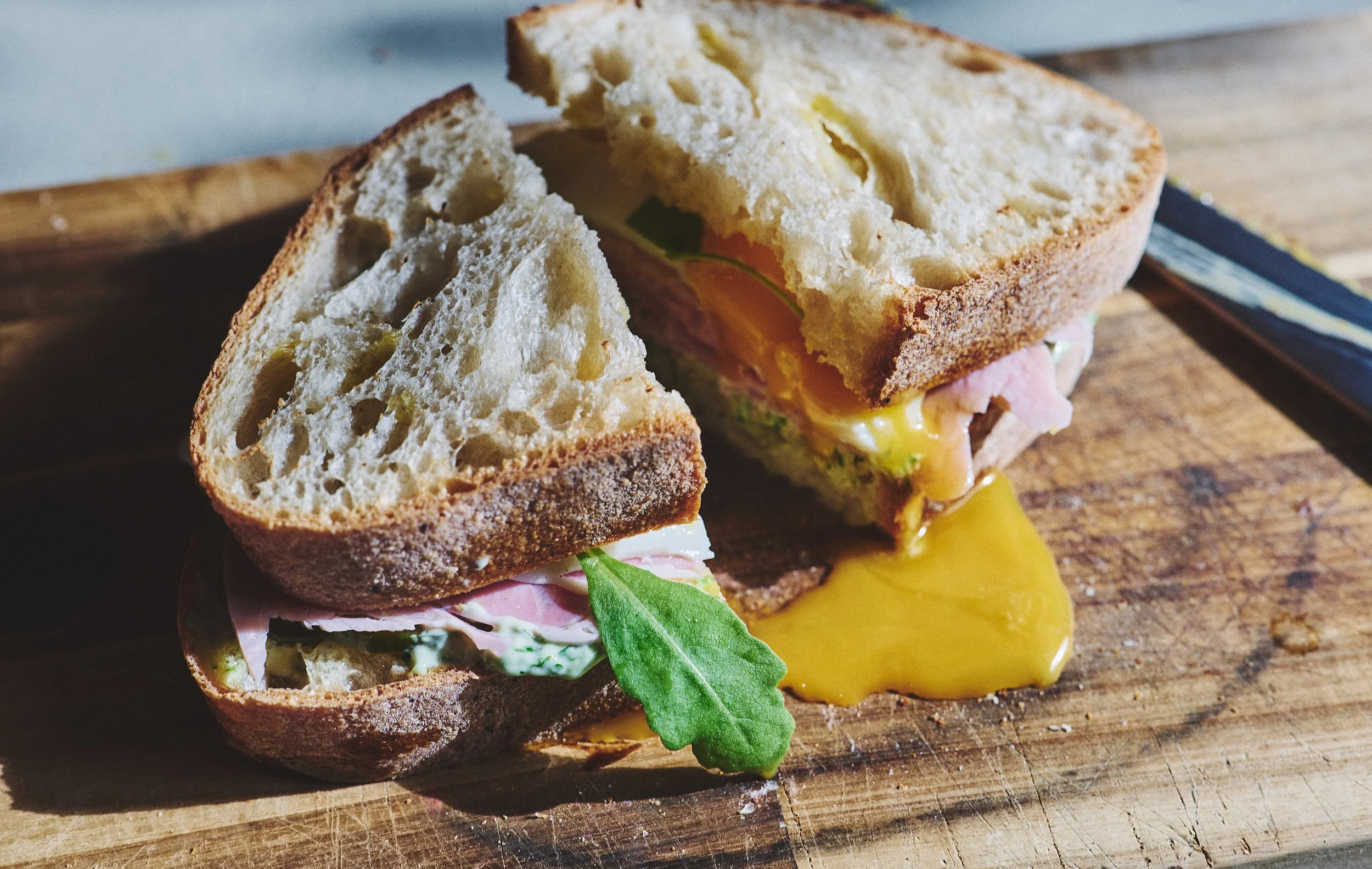 A sliced sandwich on a wooden cutting board with egg, lettuce, and ham inside, with some egg spilling out and a green leaf on the inside.