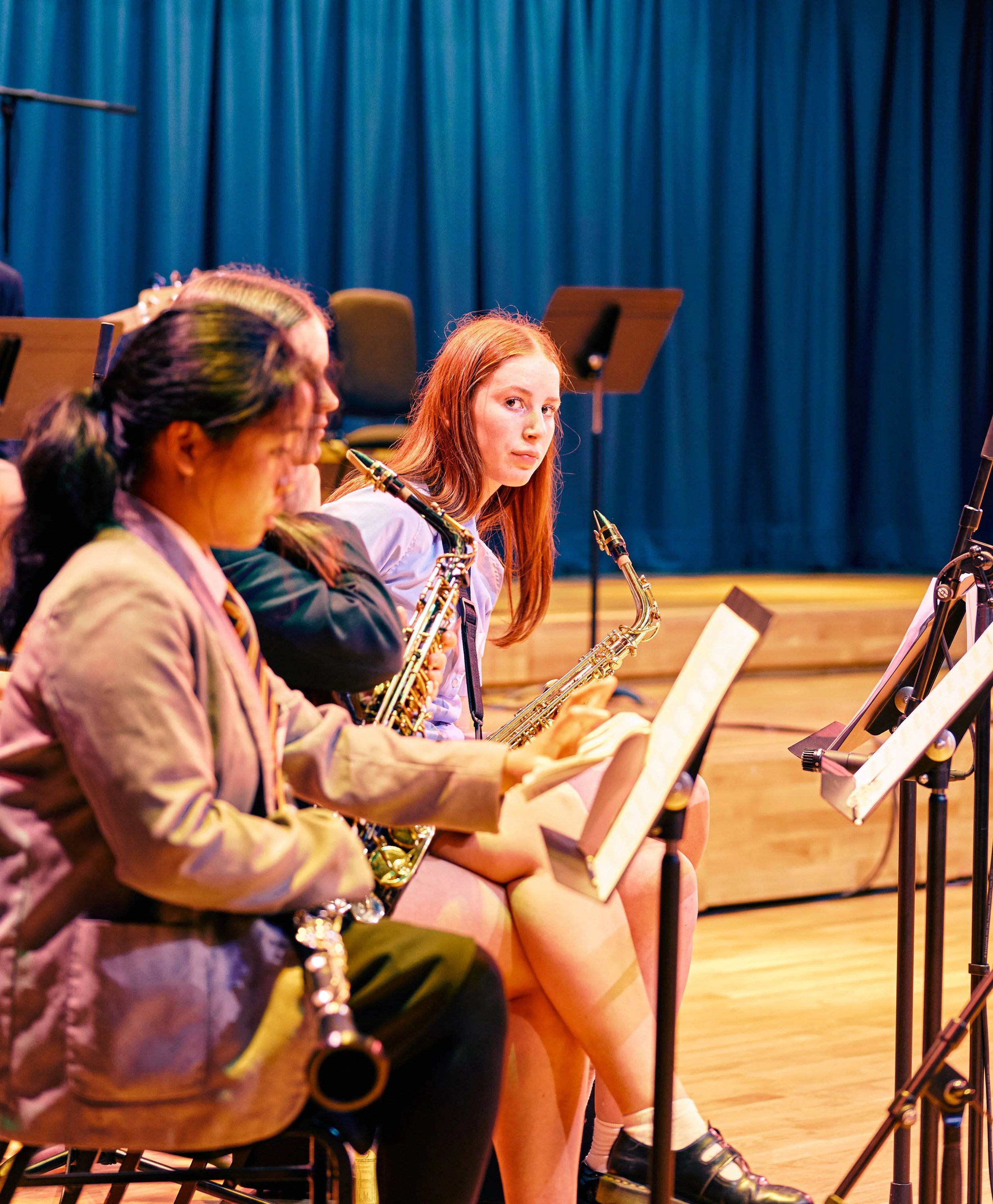 Young female orchestra musician with long red hair looking directly at the camera while seated with others on stage, holding a saxophone, in a concert hall with blue curtains.