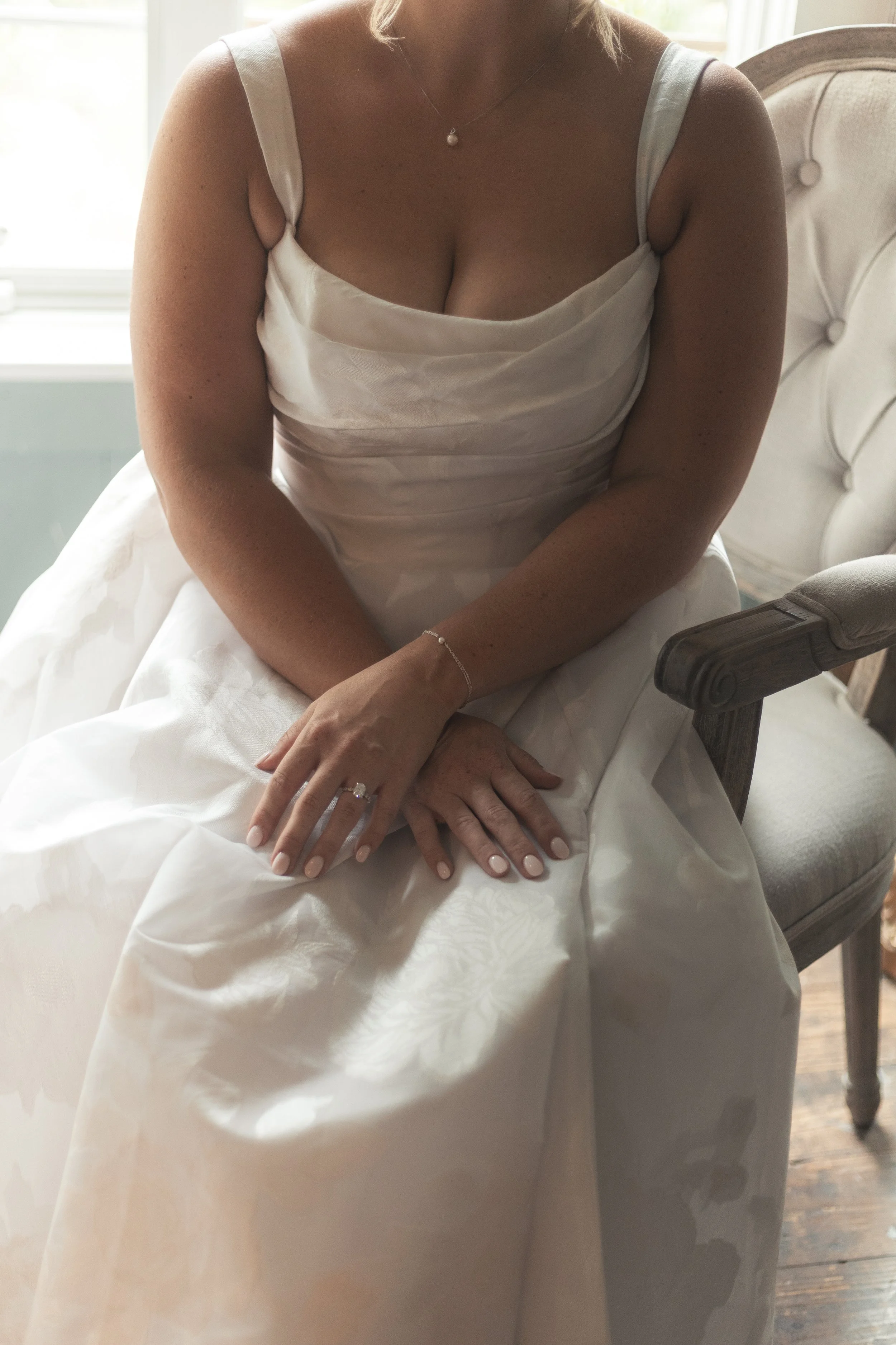 A woman sitting on a vintage armchair in a white wedding dress, with her hands resting on her lap, wearing rings and a delicate bracelet, near a window with natural light.