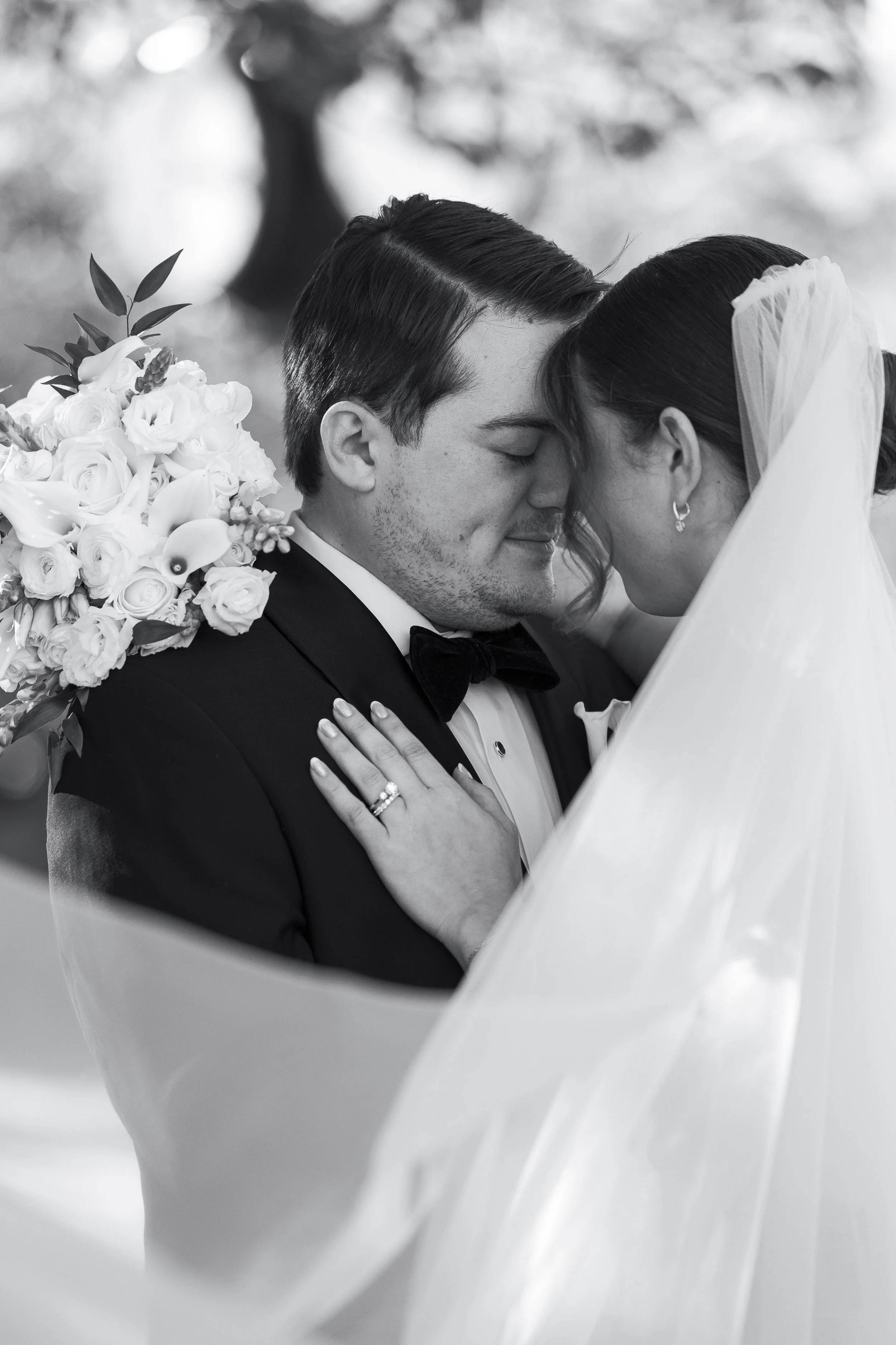 Black and white photo of a bride and groom close together, forehead to forehead, with eyes closed, outdoors. The bride wears a veil and earrings, and the groom wears a tuxedo and bow tie. The bride's hand with an engagement ring rests on the groom's 