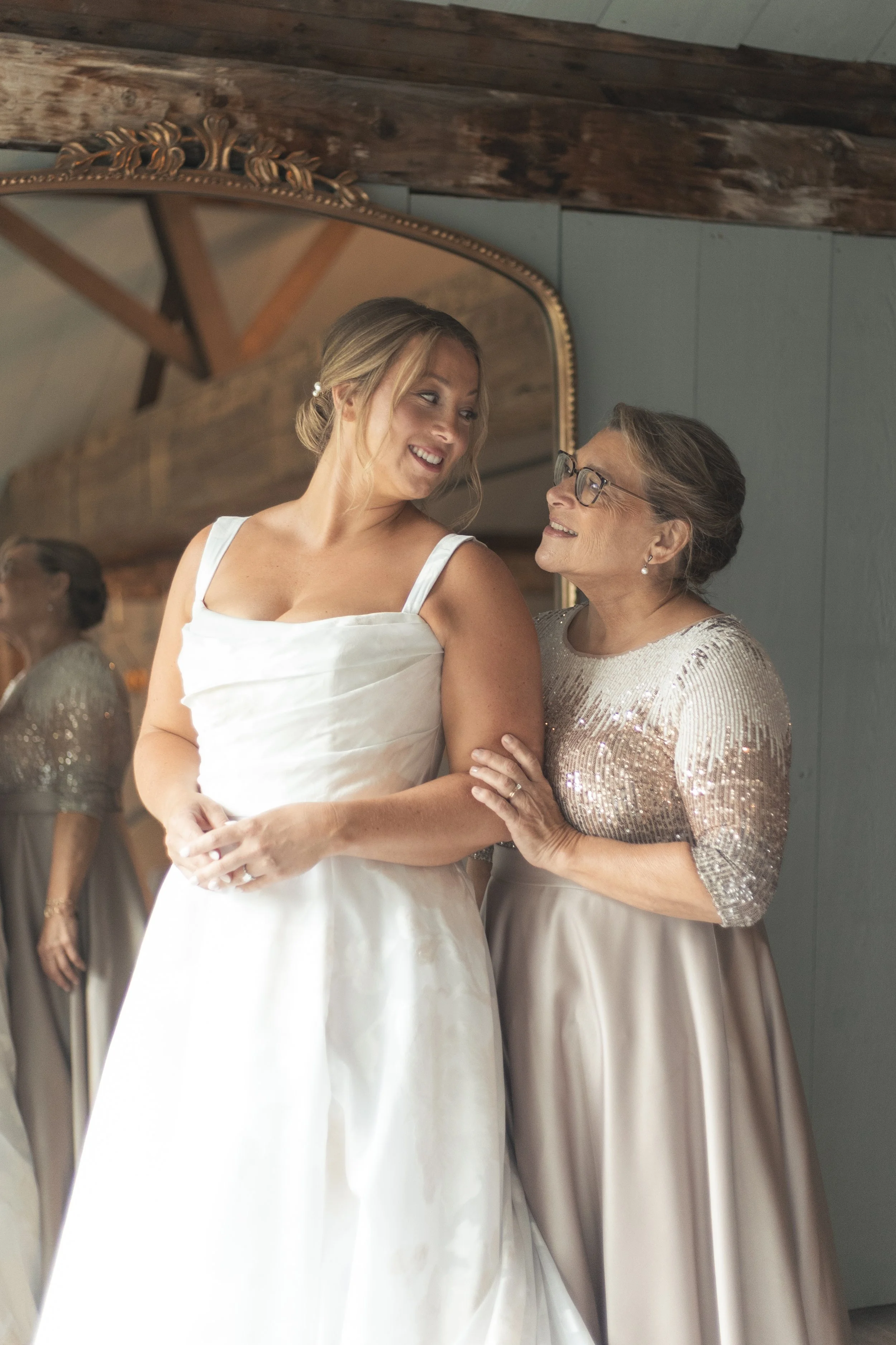 A bride in a white wedding dress smiling at her mother, who is wearing a sparkly top and a satin skirt, in a room with wooden beams and a large mirror.
