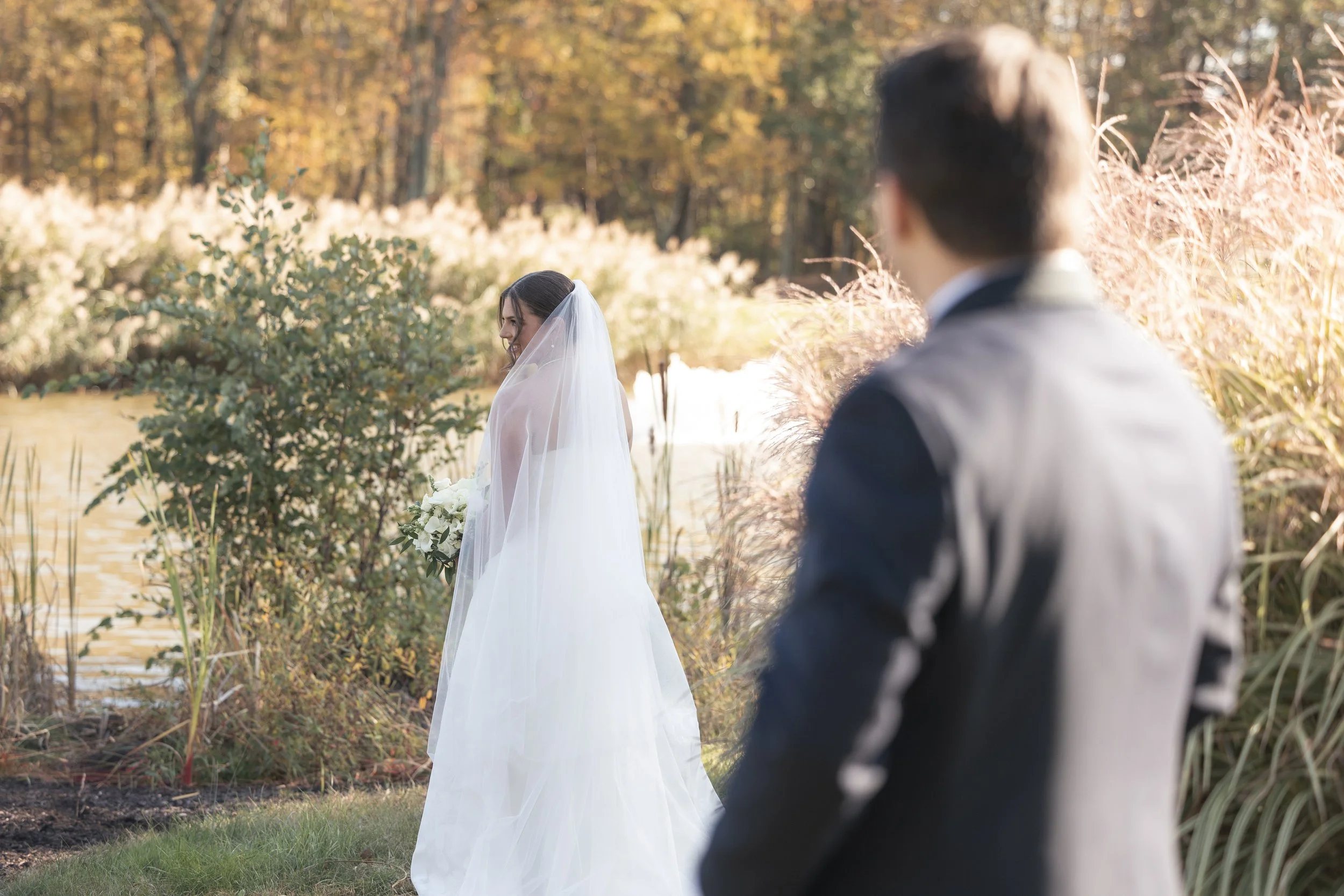 Wedding couple standing outdoors near a lake in a wooded area with fall foliage. The bride is in a white wedding dress and veil, holding a bouquet of white flowers. The groom is in a dark suit, standing behind and to the right of the bride, looking a
