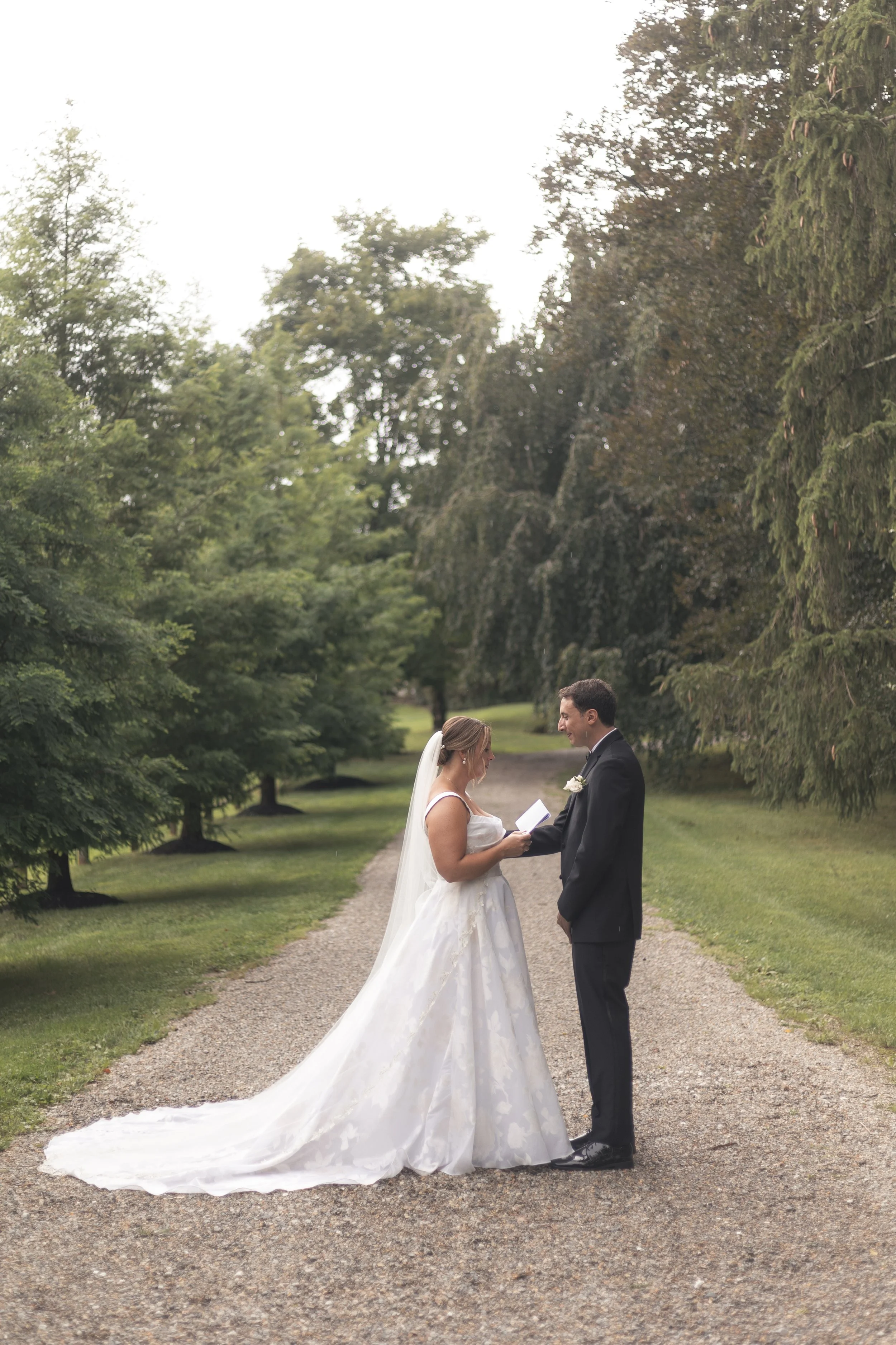 Bride and groom exchanging vows on a gravel path surrounded by lush green trees.