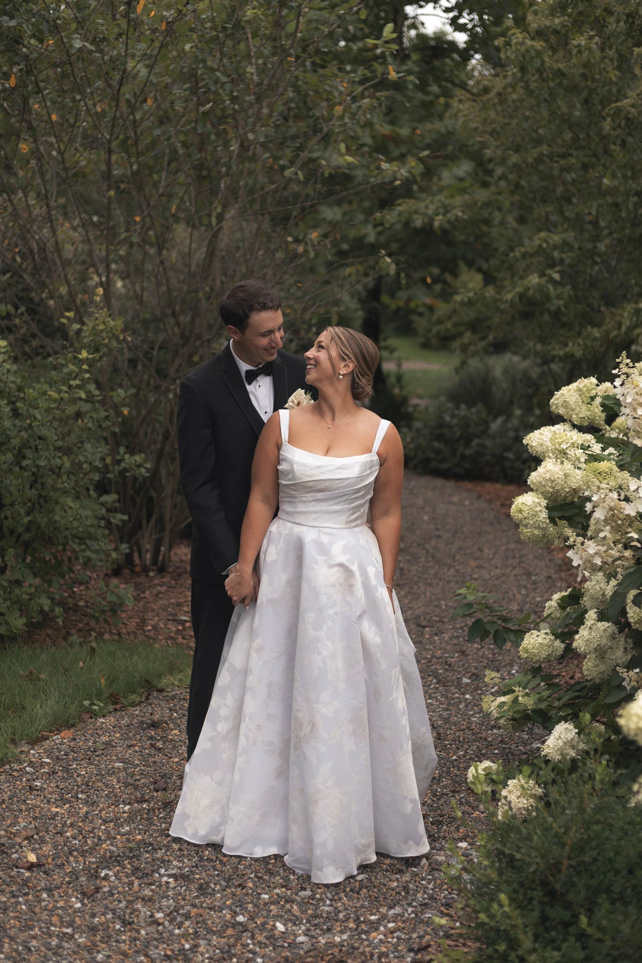 A bride and groom stand together on a garden pathway, smiling at each other. The bride wears a white wedding gown with floral patterns and the groom is dressed in a black tuxedo with a bow tie. Surrounding them are green trees and blooming white hydr