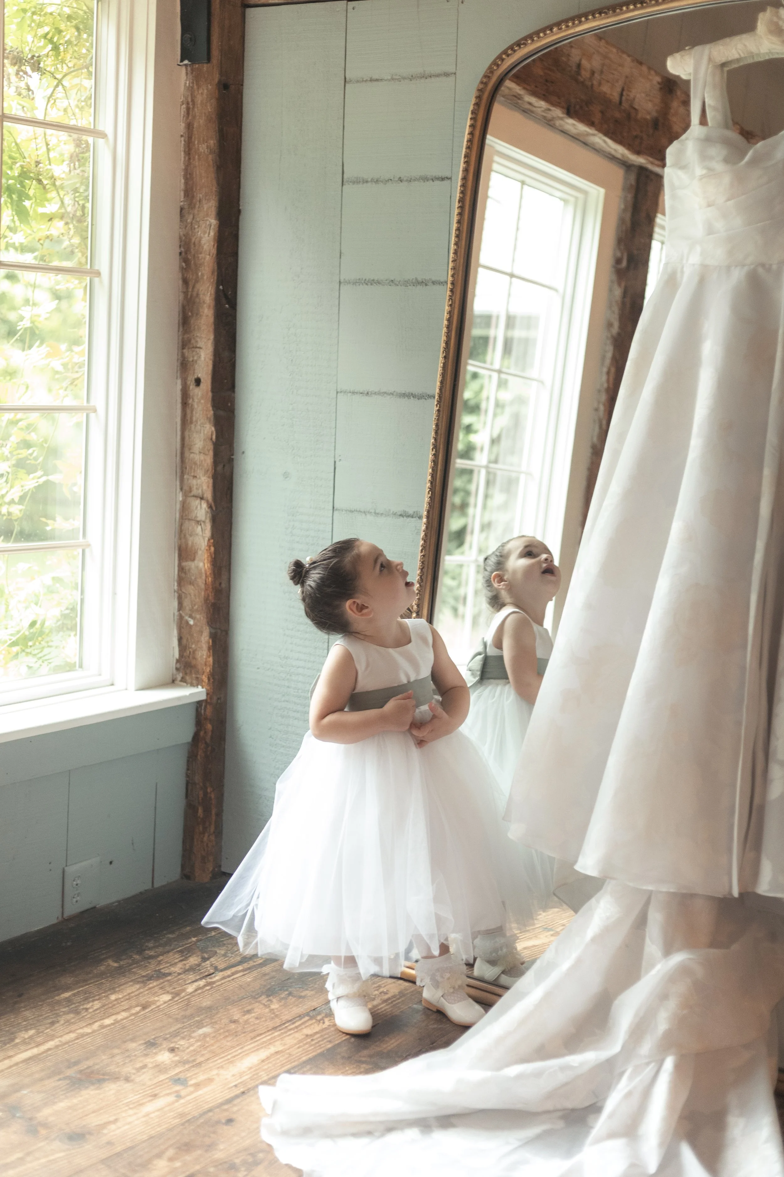 A young girl in a white dress with a gray sash looking at a wedding dress in a mirror, with trees visible outside the window.