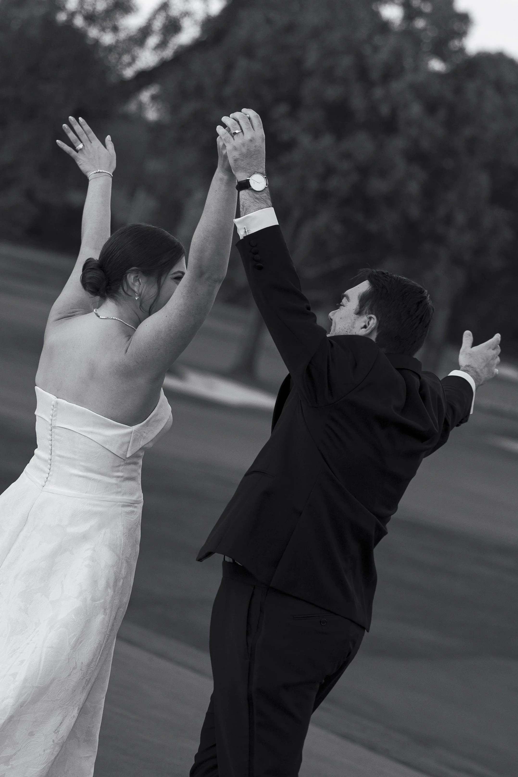 A bride and groom are dancing outdoors with their hands raised, smiling at each other. The image is in black and white.