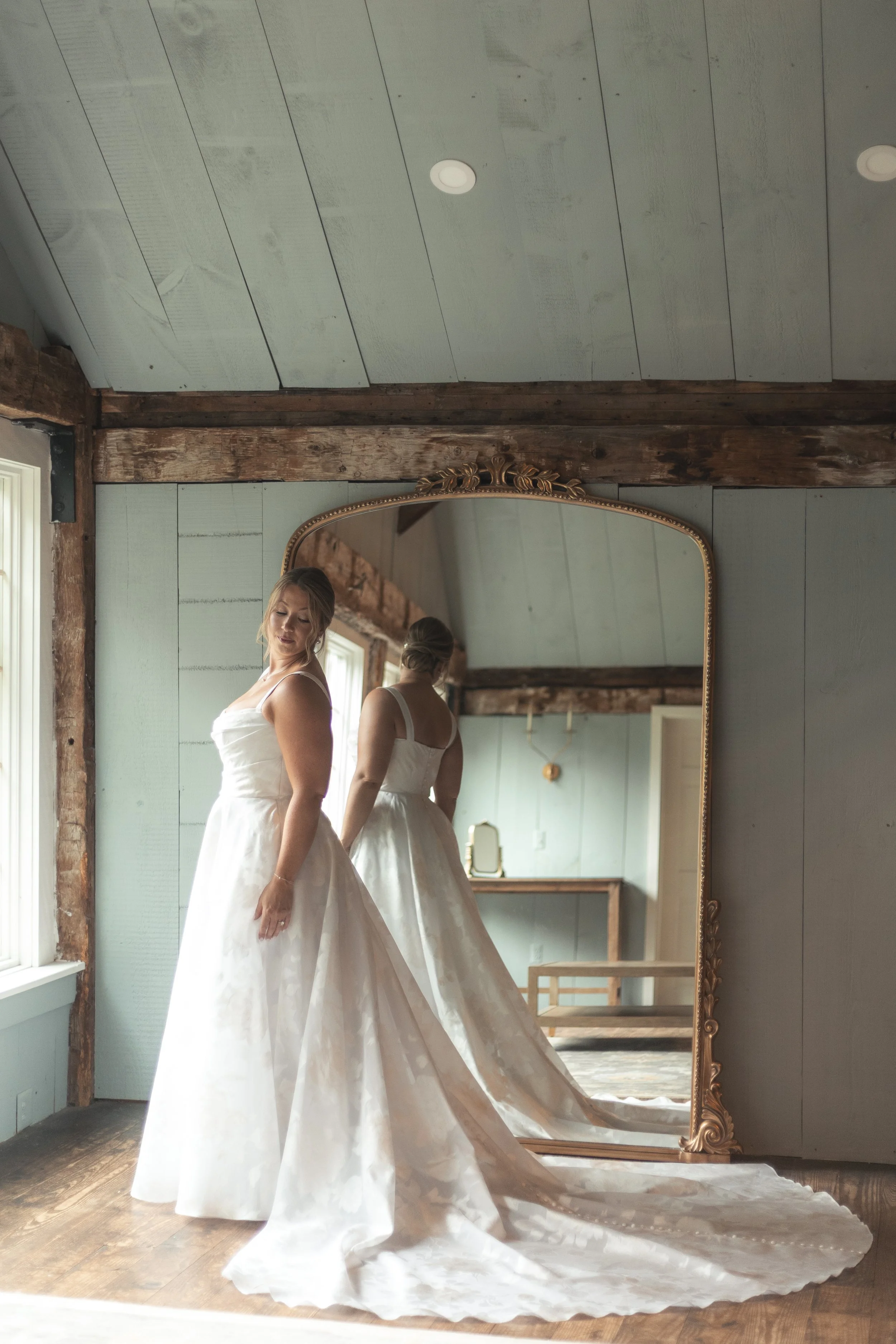 A bride in a white wedding dress looking at herself in a large ornate mirror in a rustic room with wooden beams and light blue walls.