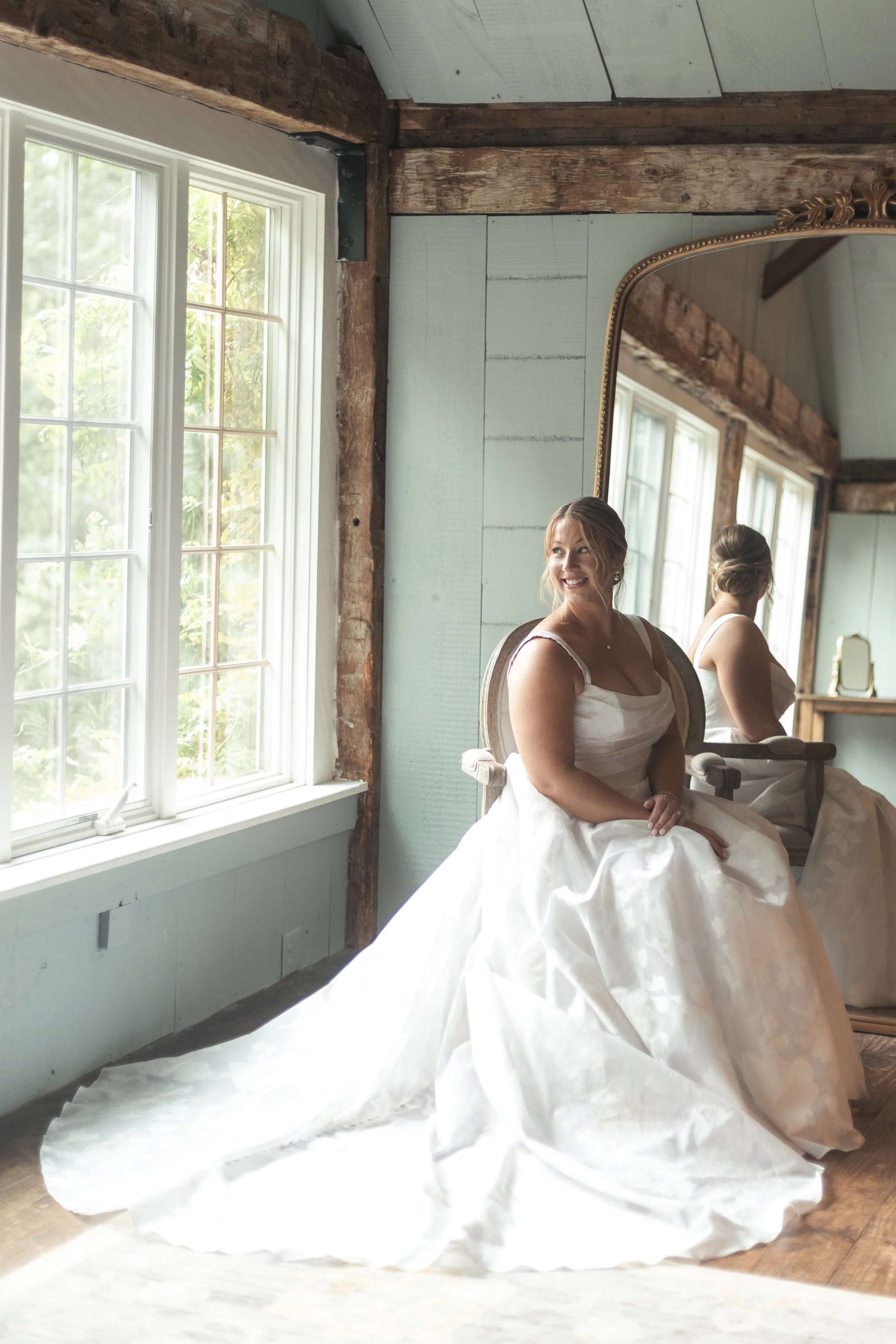 A woman in a white wedding dress sitting on an antique chair in front of a large mirror, smiling, with sunlight coming through a window beside her.