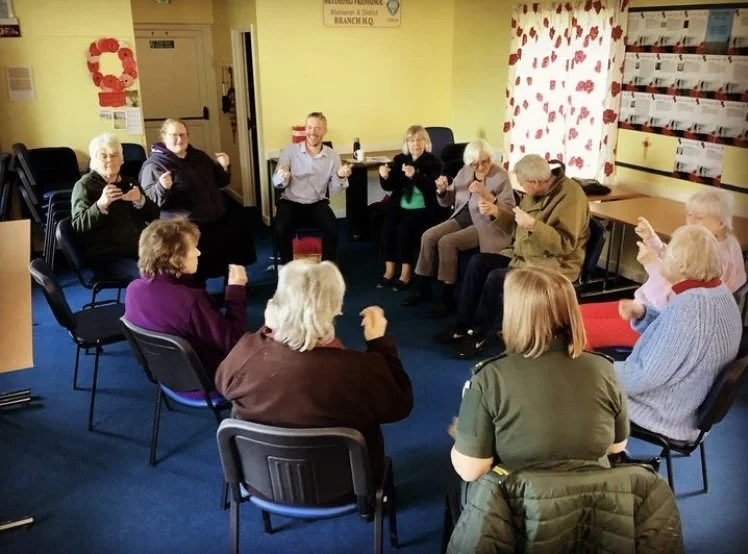 A group of elderly people sitting in a circle in a room, participating in a group activity or game, some are smiling and holding small objects or cards.
