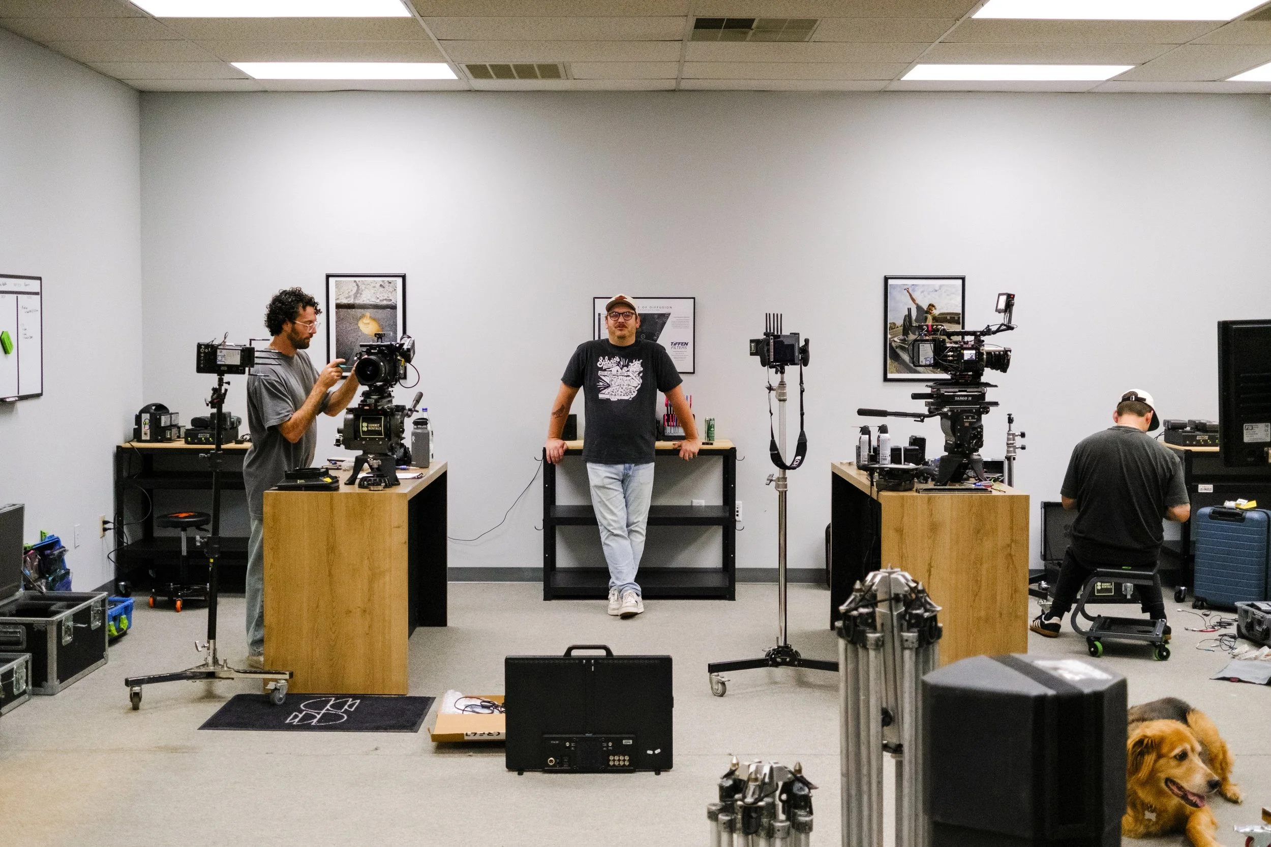 A filming studio with three men working with cameras, equipment, and monitors. One man is holding a camera, another is seated working with his equipment, and the third man is standing in the center leaning on a table. A golden retriever is lying on the floor at the right corner.