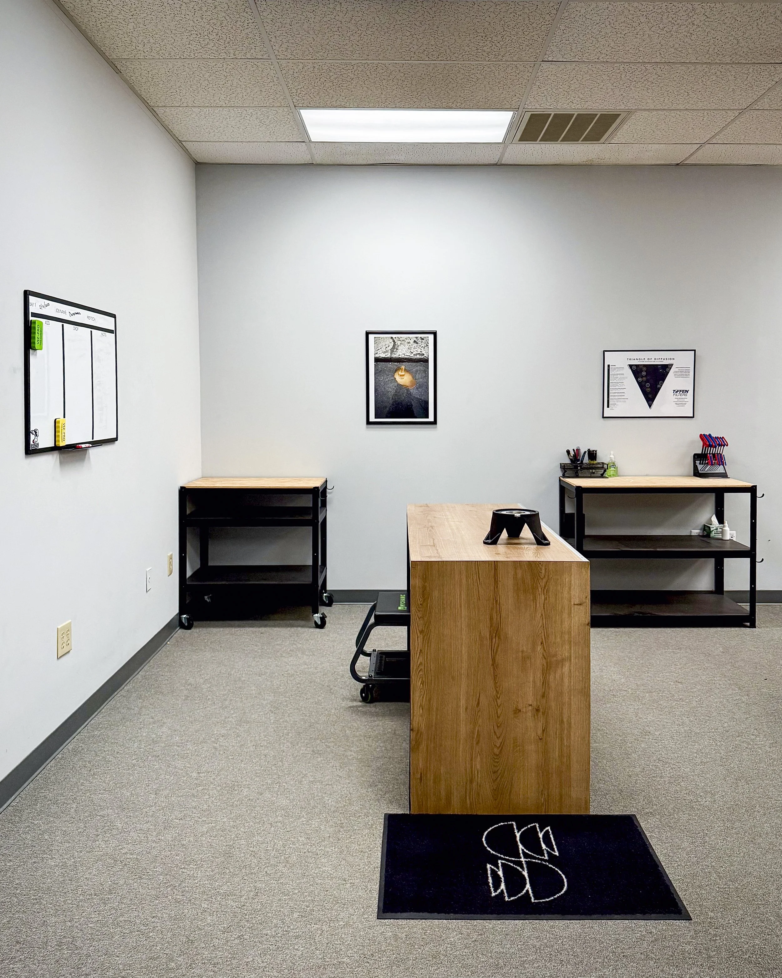Empty office space with a wooden reception desk, black rolling cart, black shelf with office supplies, framed pictures on the wall, and a whiteboard on the wall.