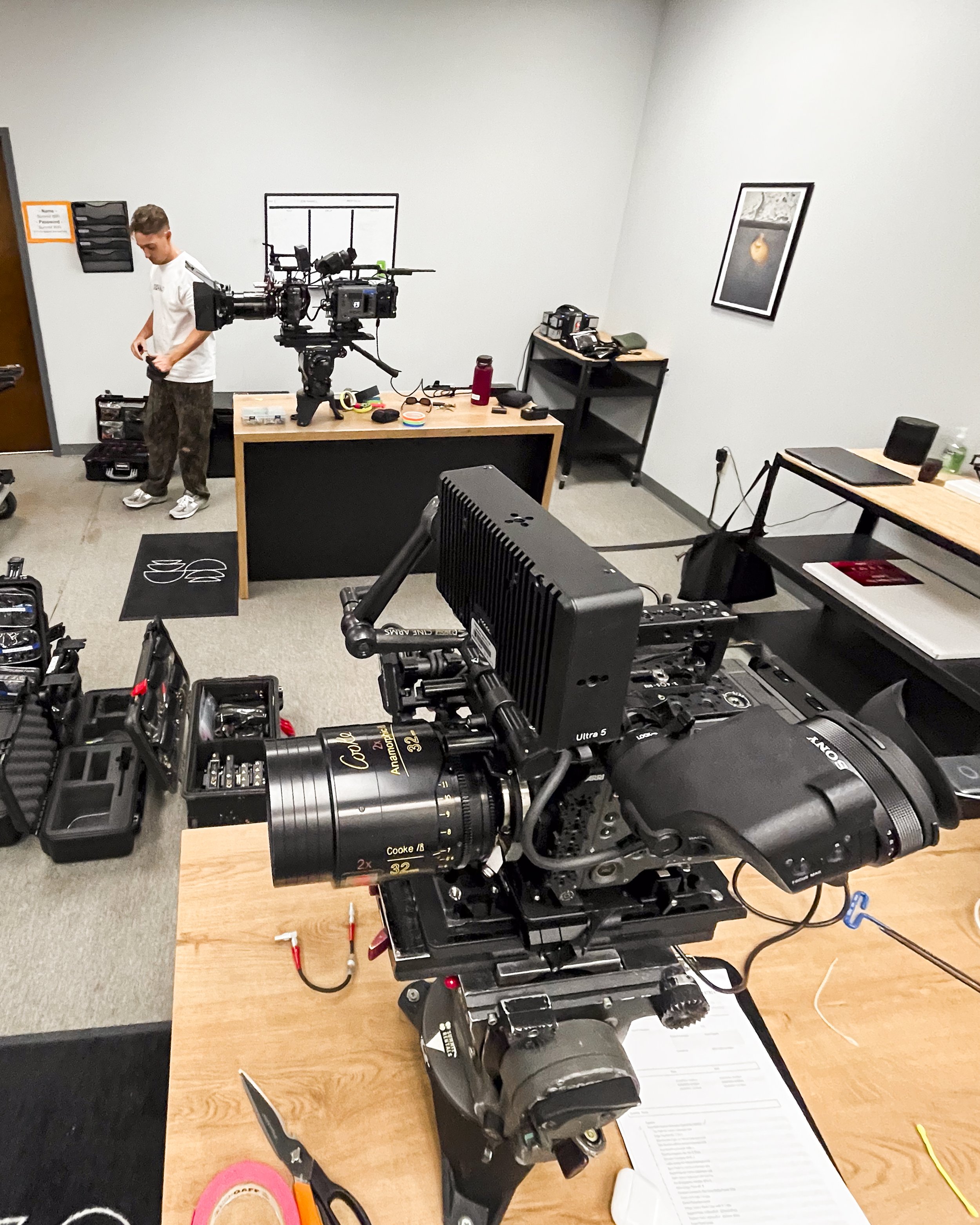 Interior of a filming studio or production room with professional camera equipment set up on a wooden table, a person adjusting equipment in the background, and various items including cables, office supplies, and framed pictures on the walls.