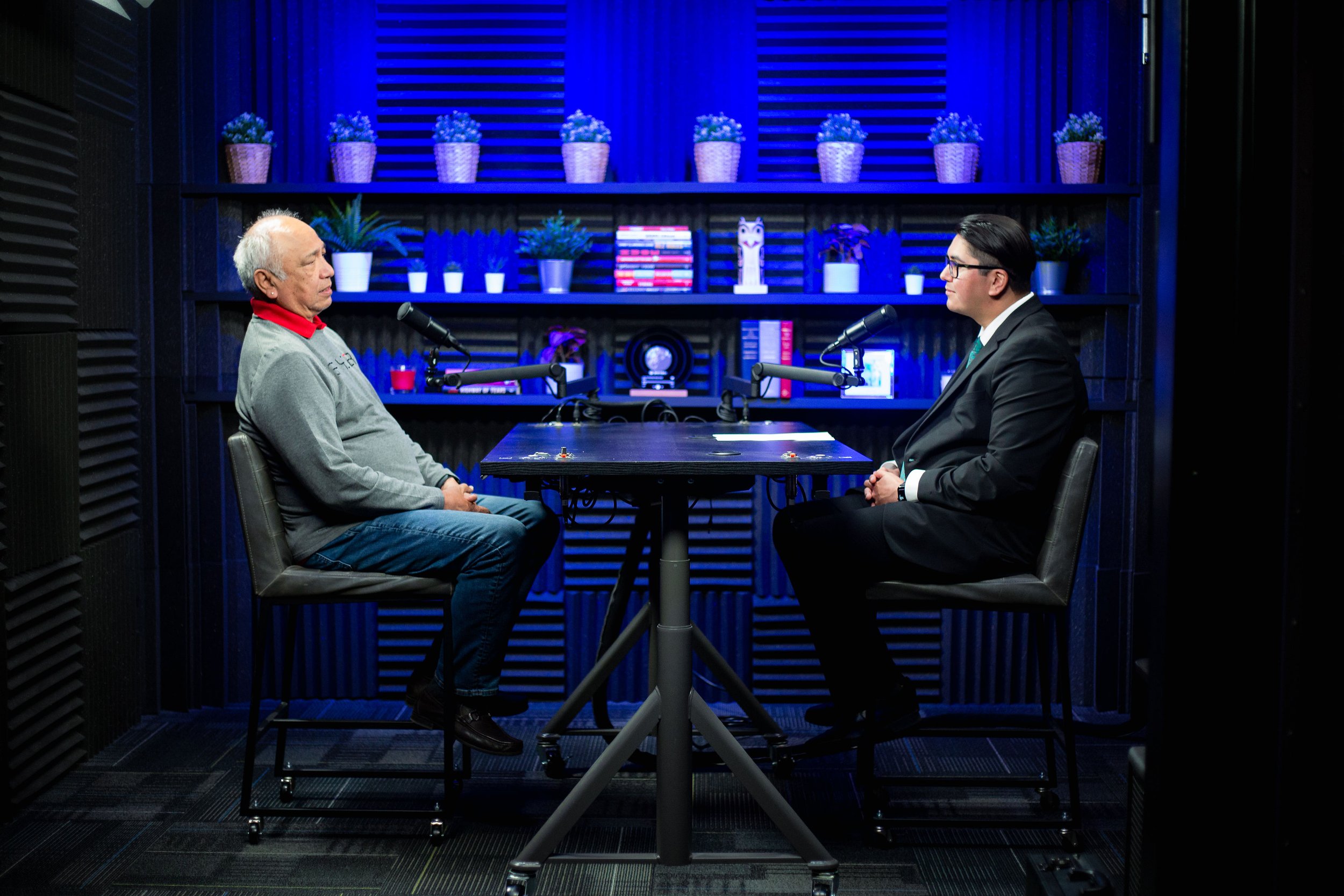 Two men sitting at a table with microphones, facing each other, in a studio with blue lighting and shelves of potted plants and books in the background.