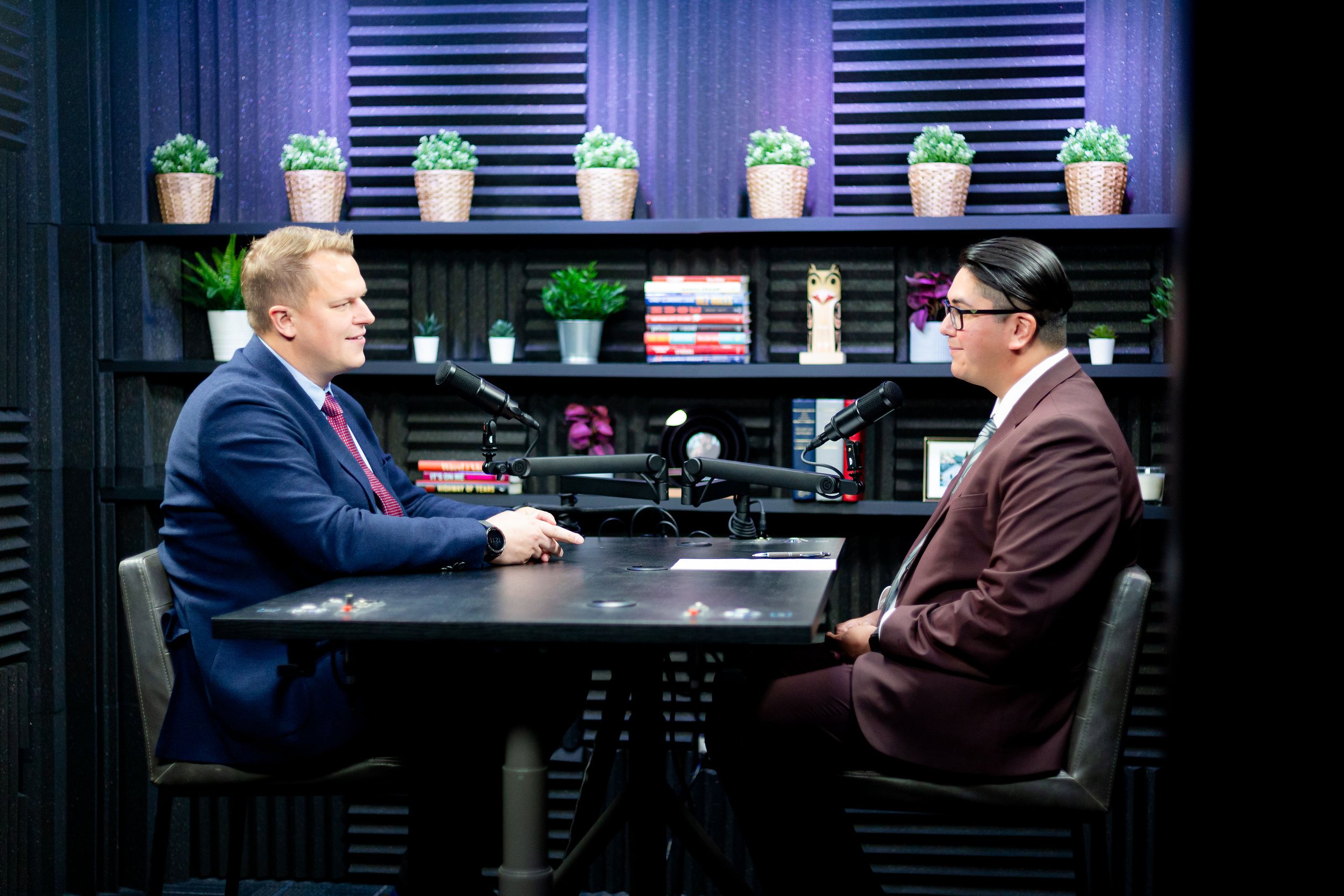 Two men in business attire having a conversation in a studio with microphones on a black table, surrounded by books and plants on shelves behind them.