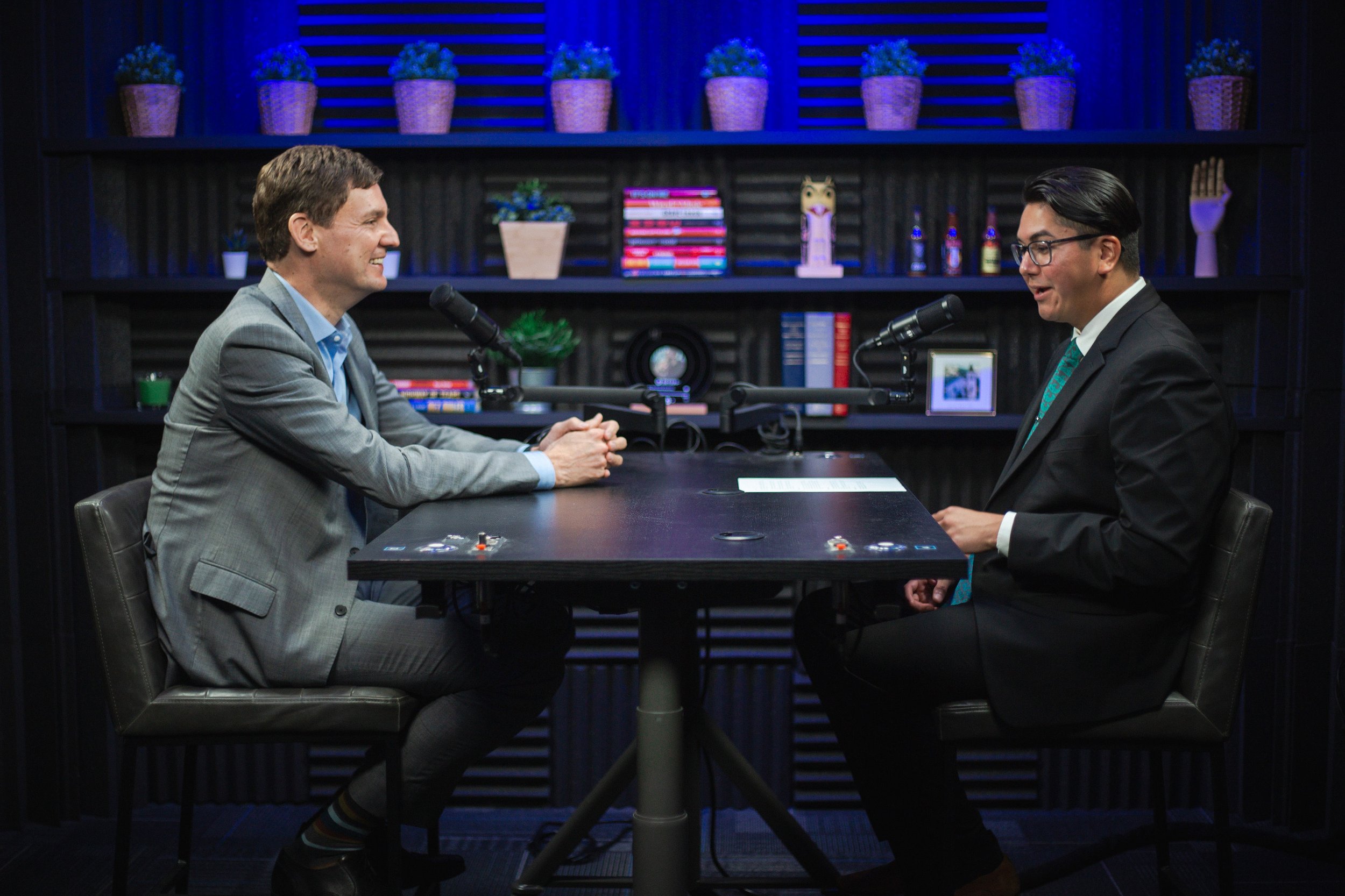 Two men in business suits are sitting at a black table with microphones, engaged in a conversation. The background features purple potted plants, books, and decorative items on shelves.