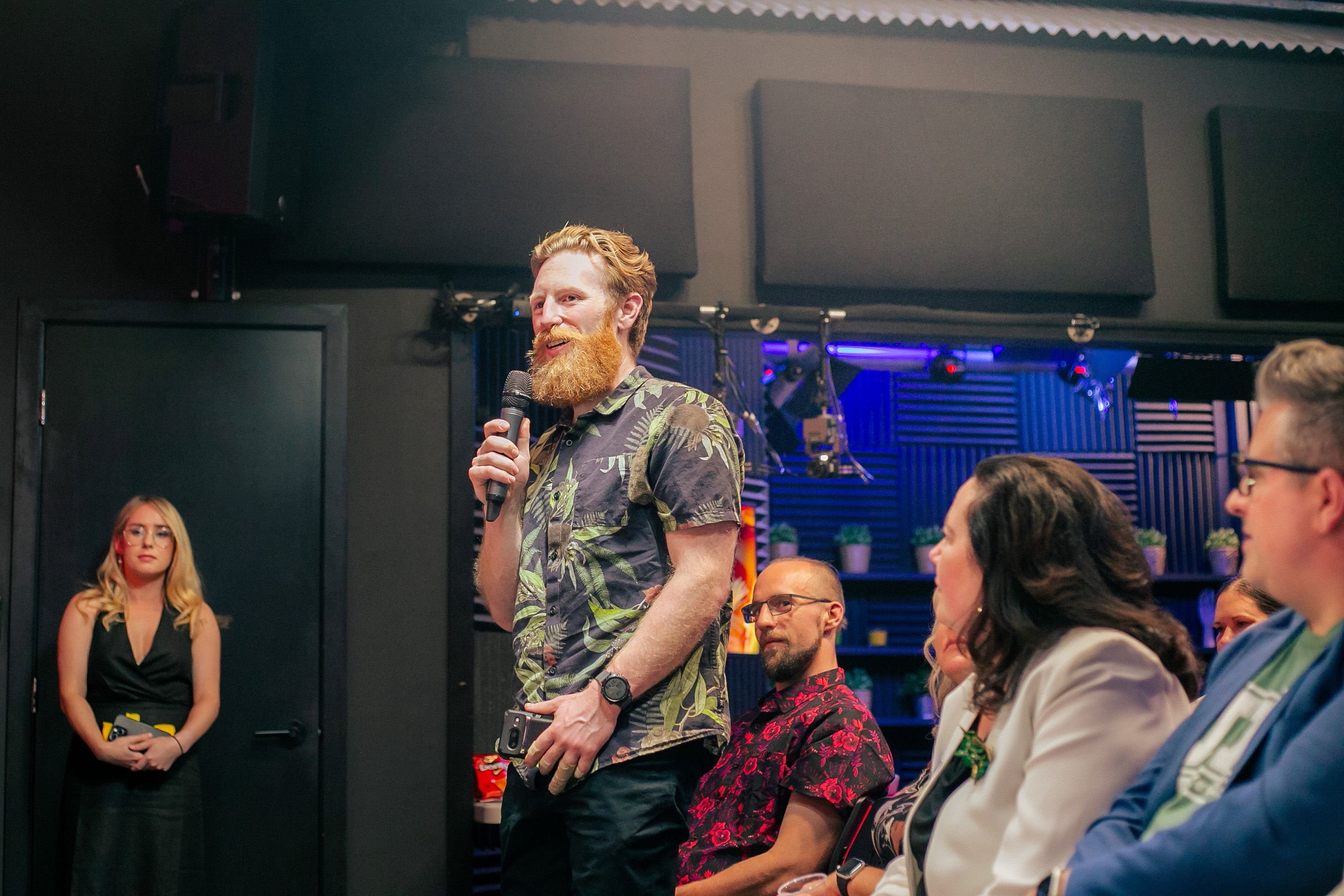 A man with red hair and beard, wearing a tropical shirt, speaking into a microphone at an indoor event, with seated audience members listening attentively.