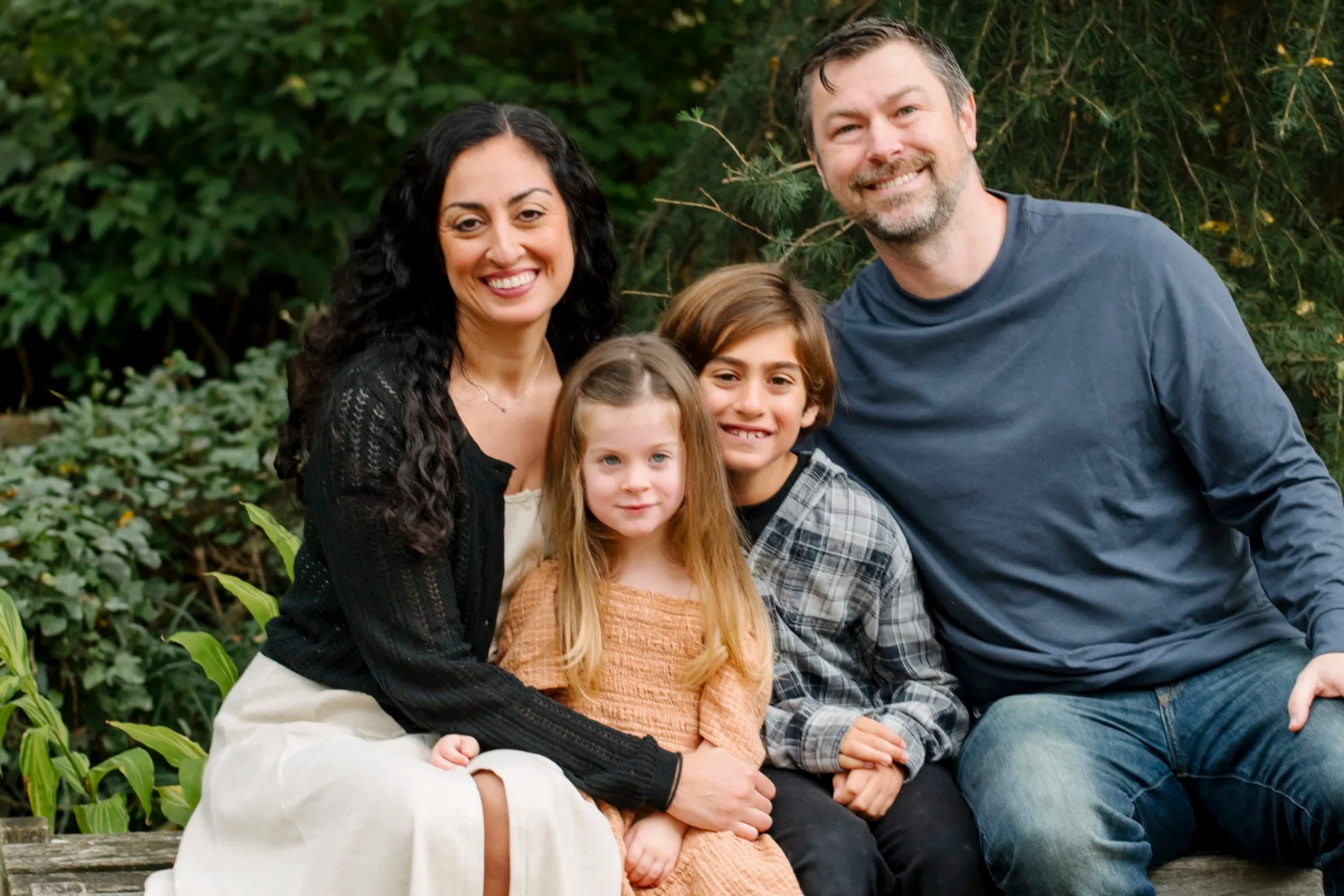 A family of four sitting outdoors on a wooden bench, surrounded by green foliage, smiling at the camera.