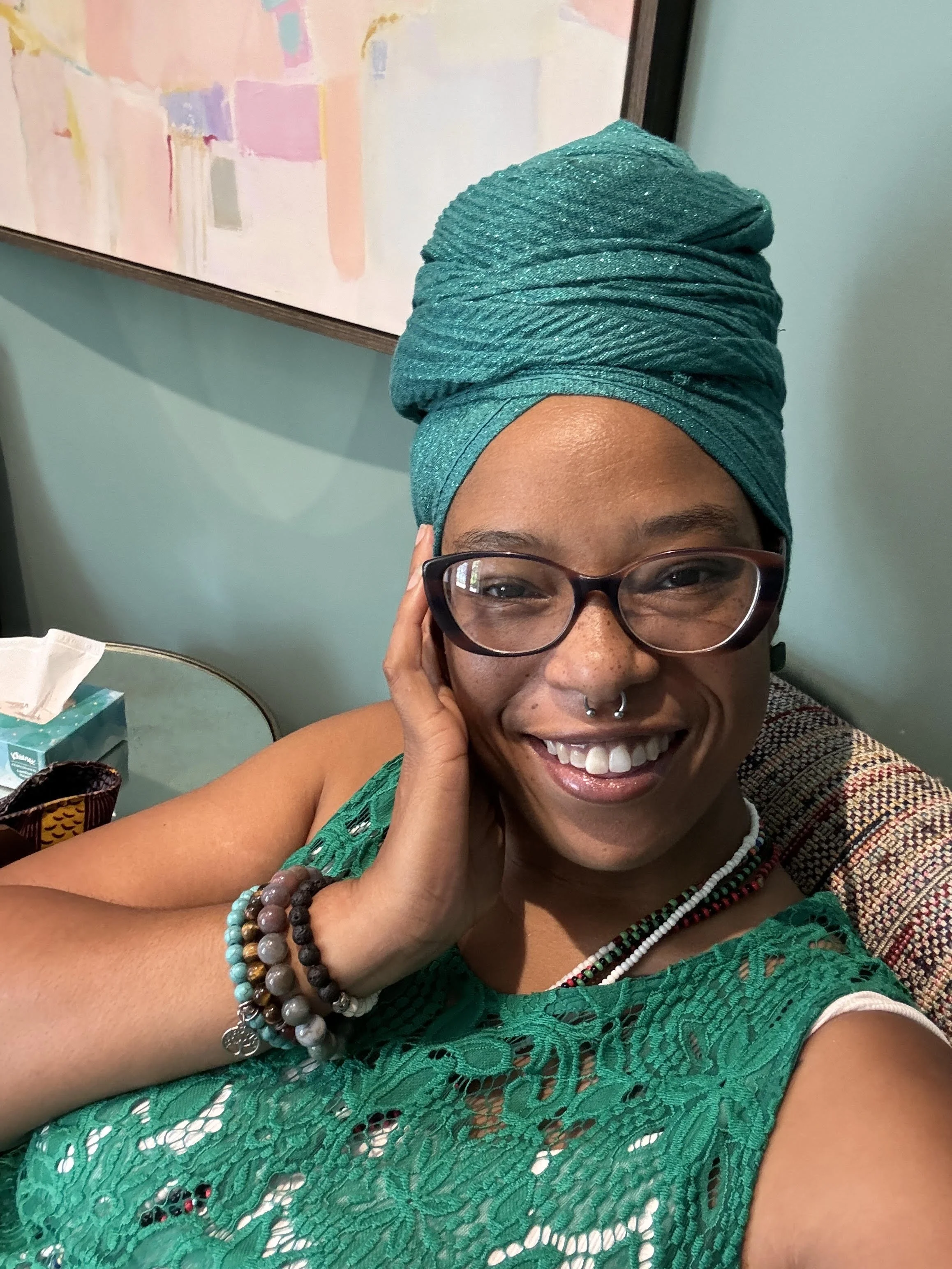 A black woman smiling, wearing glasses, a teal headwrap, a green lacy top, and beaded jewelry, sitting in a room with light green walls and artwork in the background.