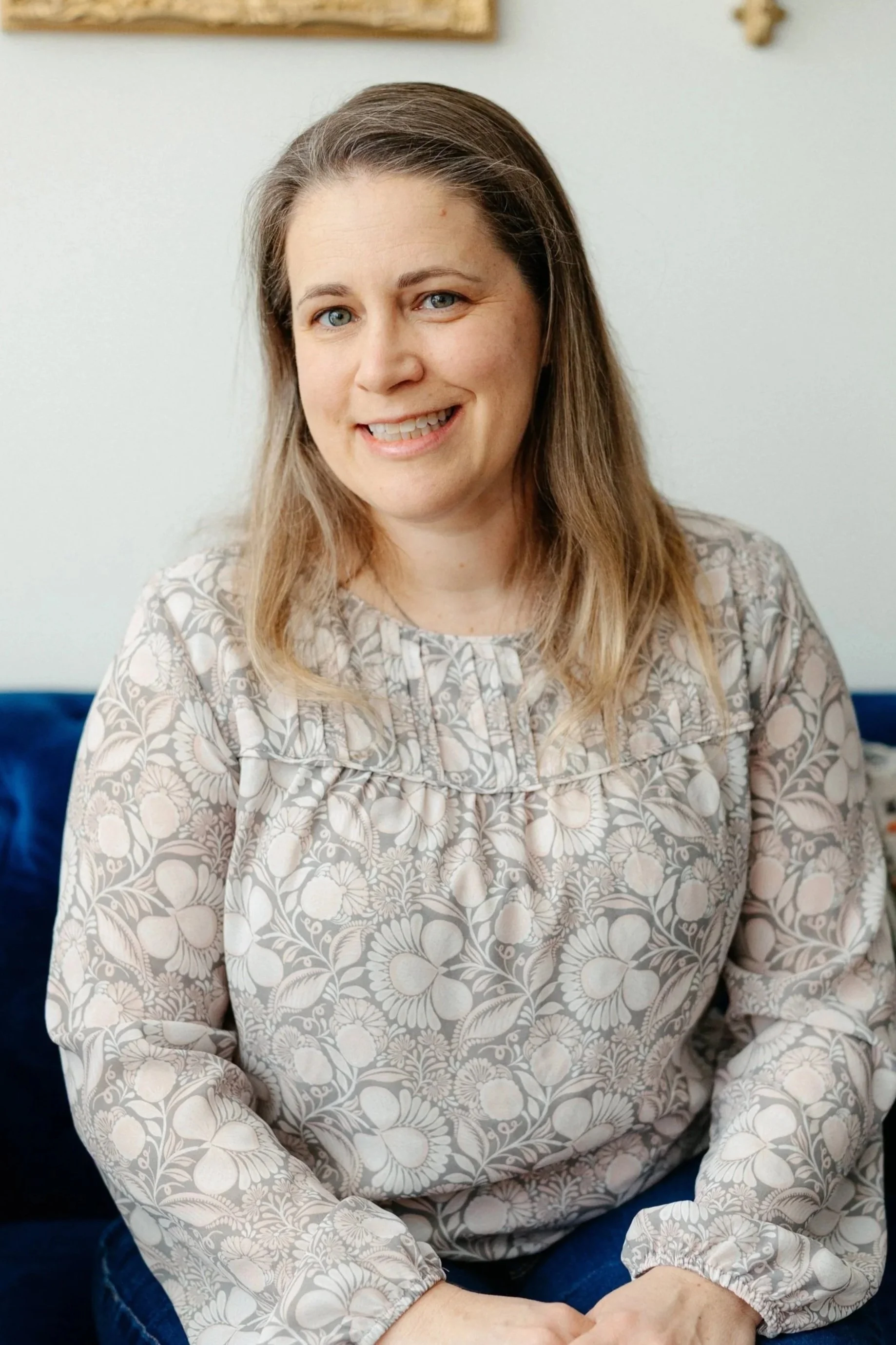 Smiling woman sitting on a blue couch while wearing a beige floral shirt
