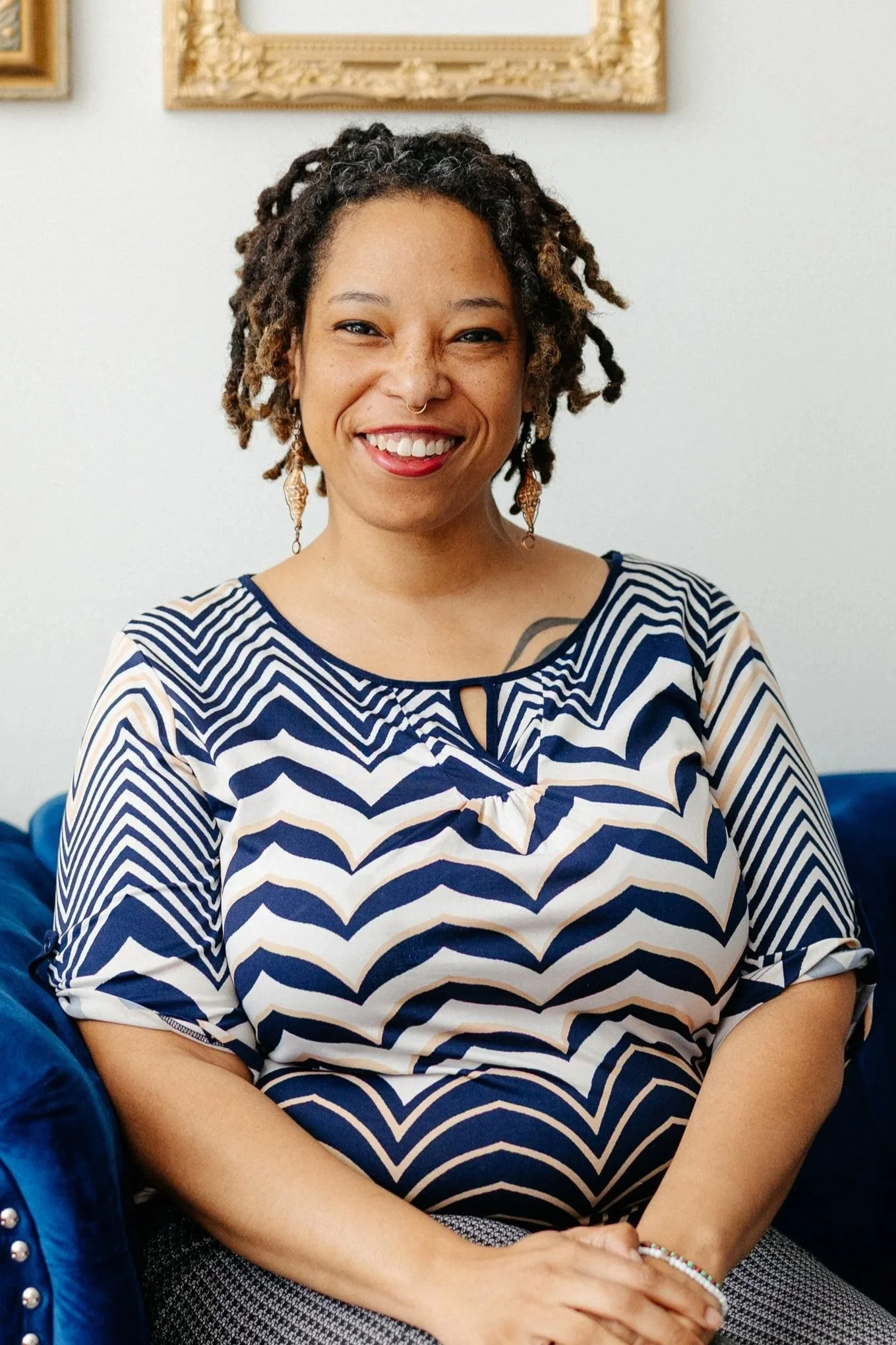 Black woman smiling while sitting on a blue couch wearing a blue and white shirt.