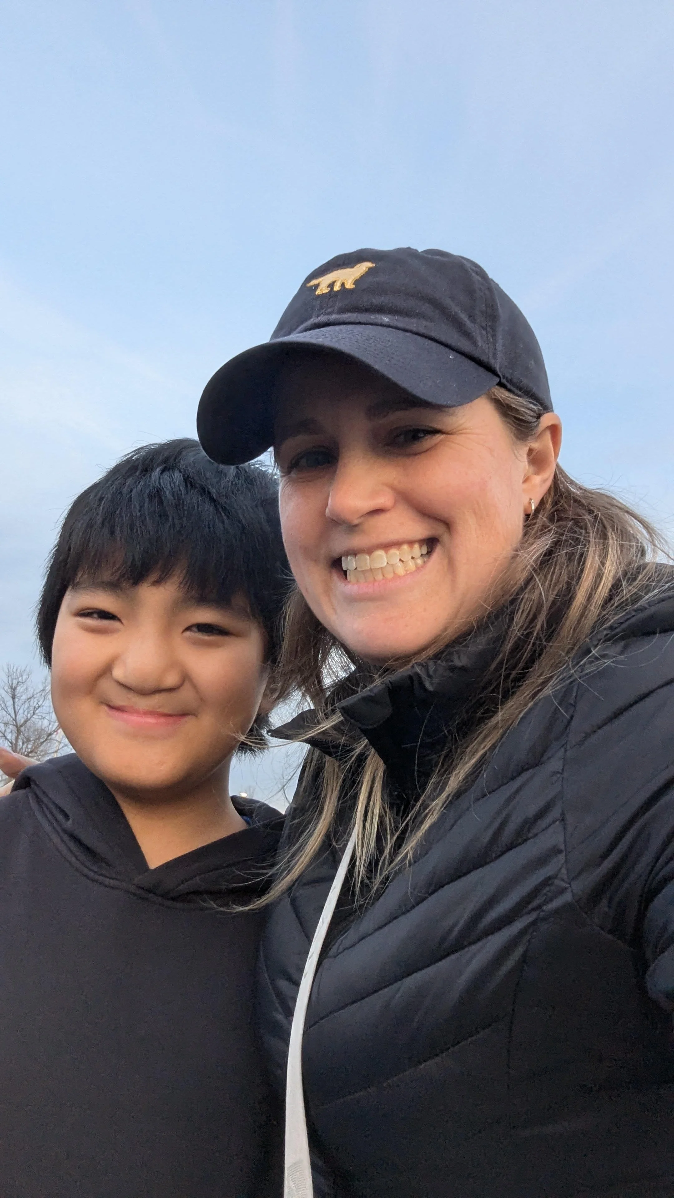 Two people smiling for a selfie outdoors, with a cloudy sky in the background. One woman wearing a baseball cap and a black jacket, and a young boy in a black hoodie.